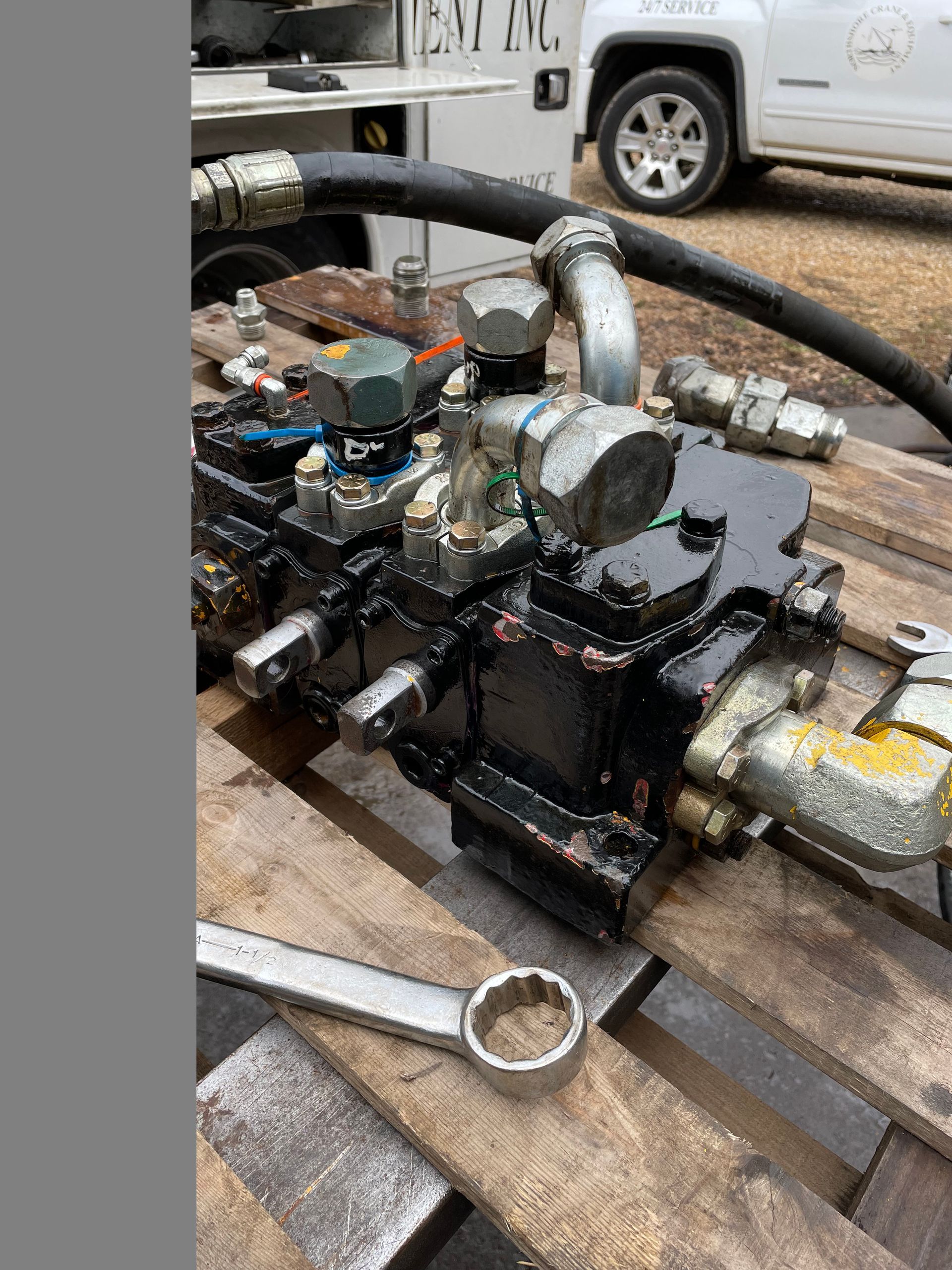 Hydraulic pump on a wooden pallet, covered in oil, with tools nearby and a truck in the background.