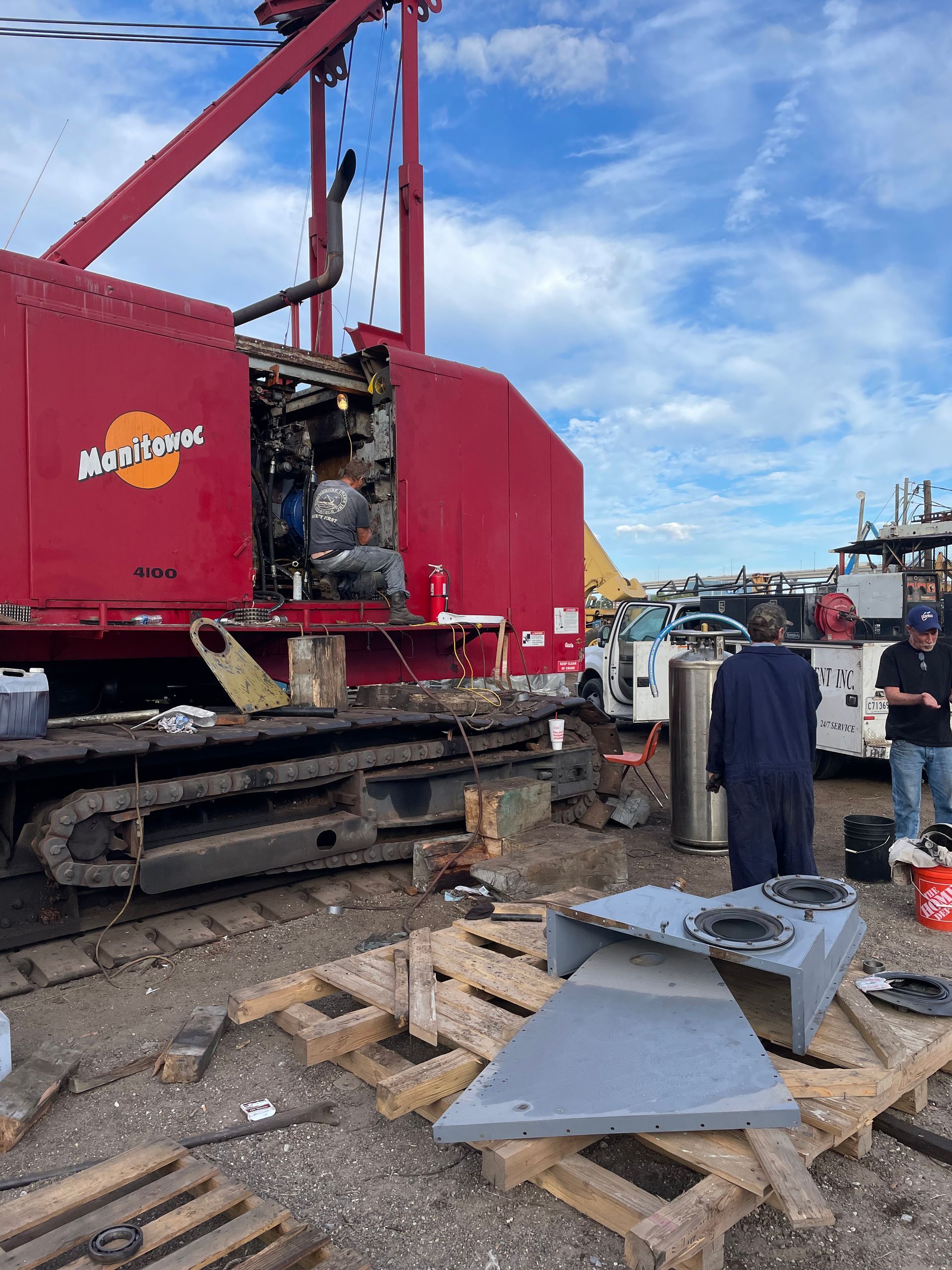 Red construction crane; workers near equipment on a paved lot. Blue sky.