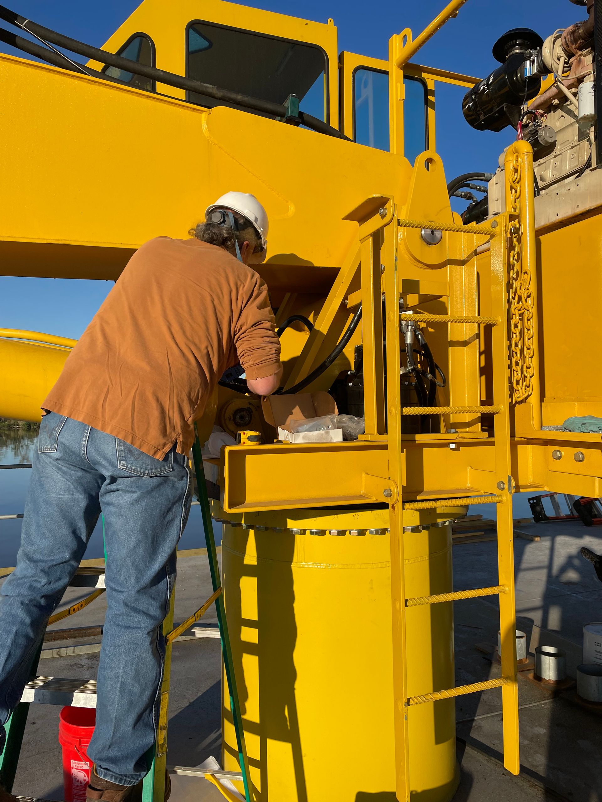 Person in jeans and brown shirt working on yellow machinery, wearing a hard hat.