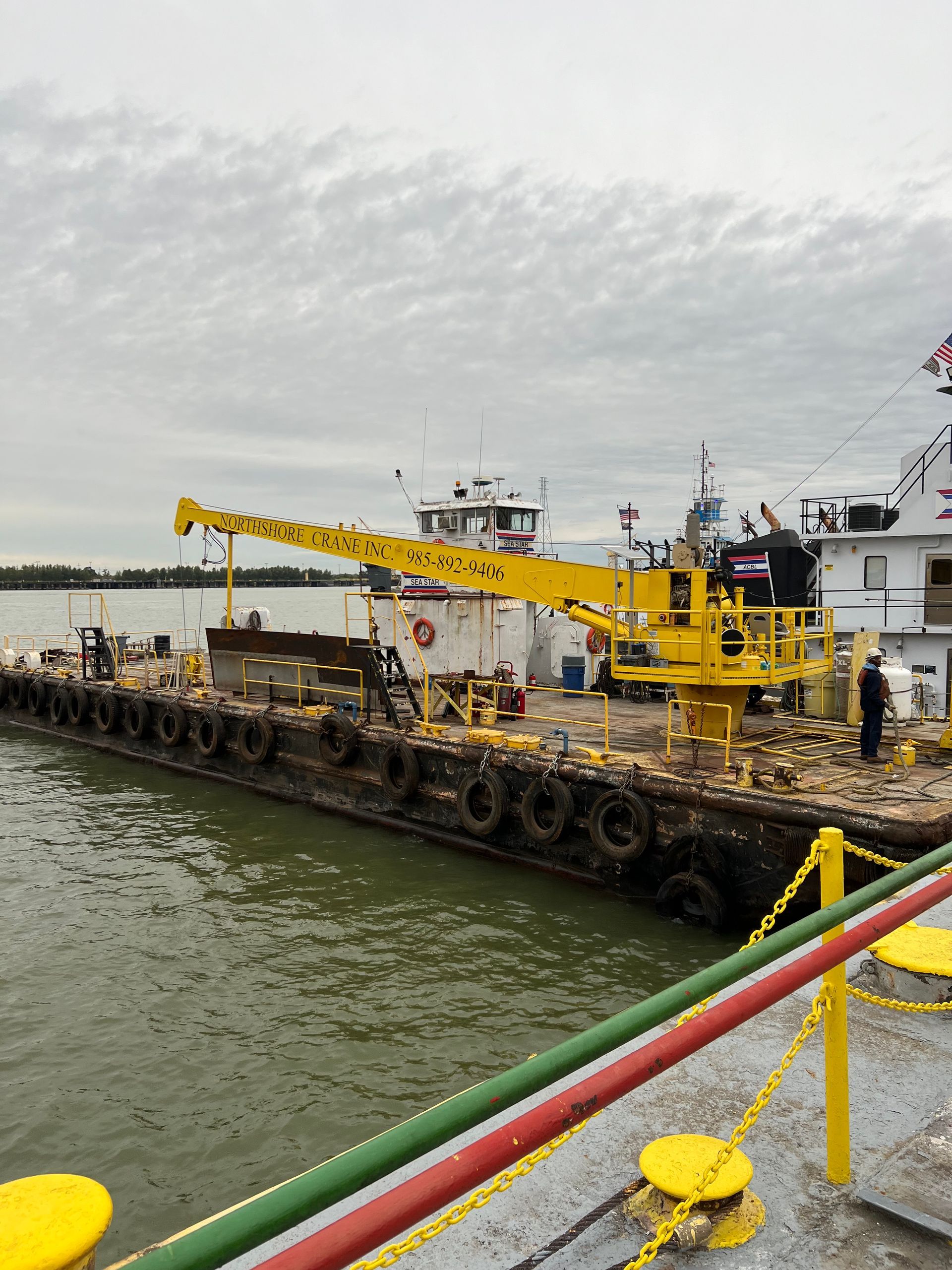Yellow crane on a dark barge by a river; overcast sky.