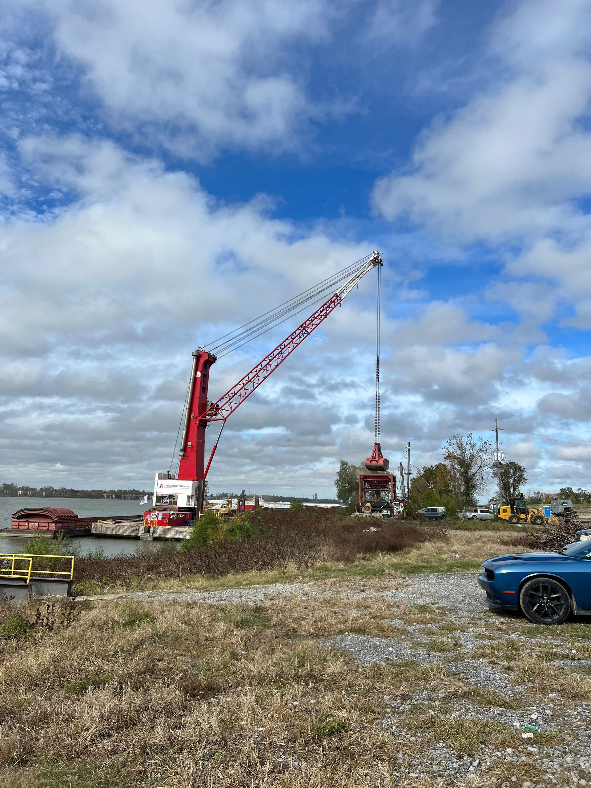 Large red and white crane on barge in river, blue car parked nearby. Cloudy sky.
