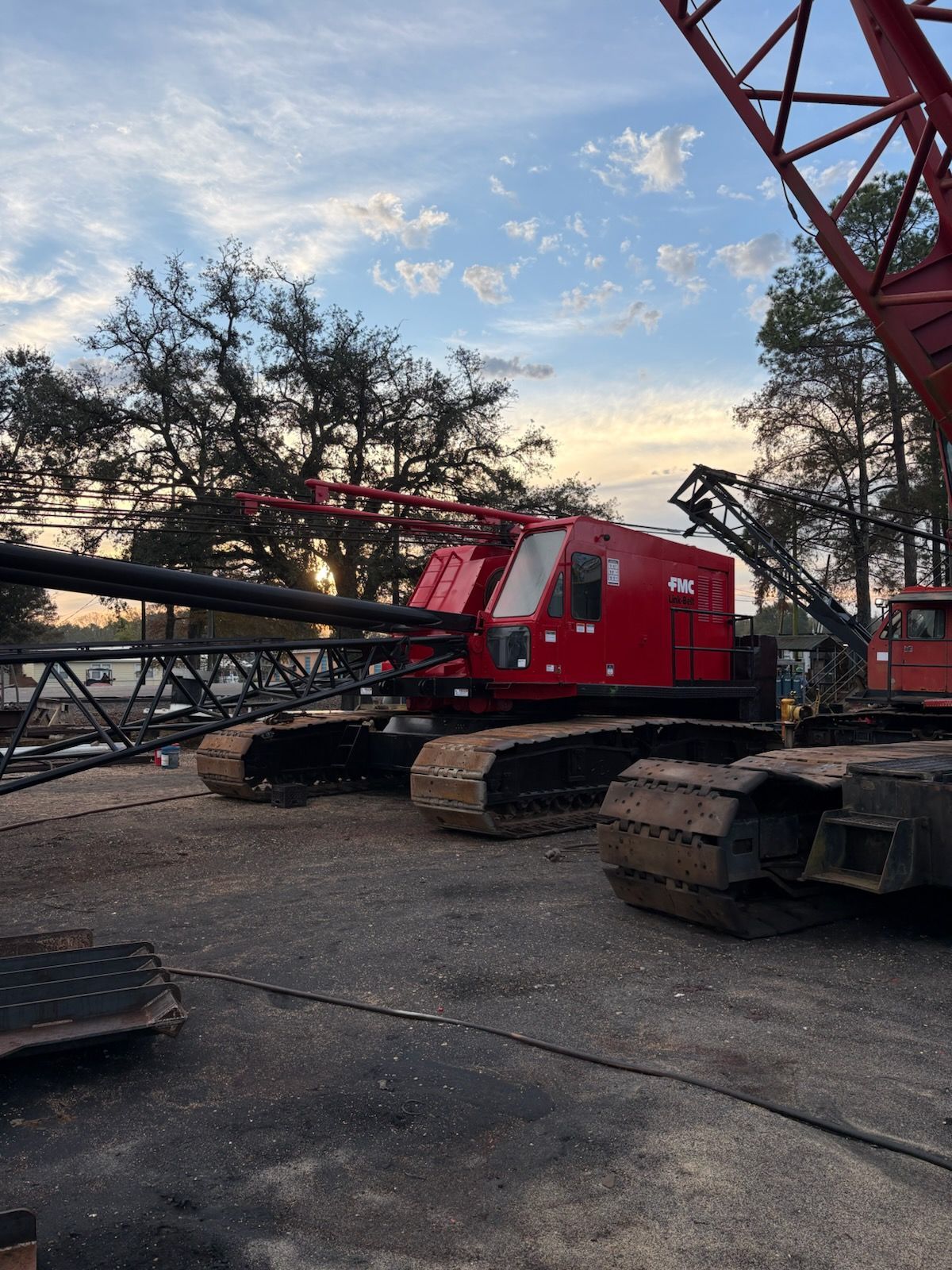 Red crawler crane on a worksite; black boom and tracks. Setting is outdoors with trees.