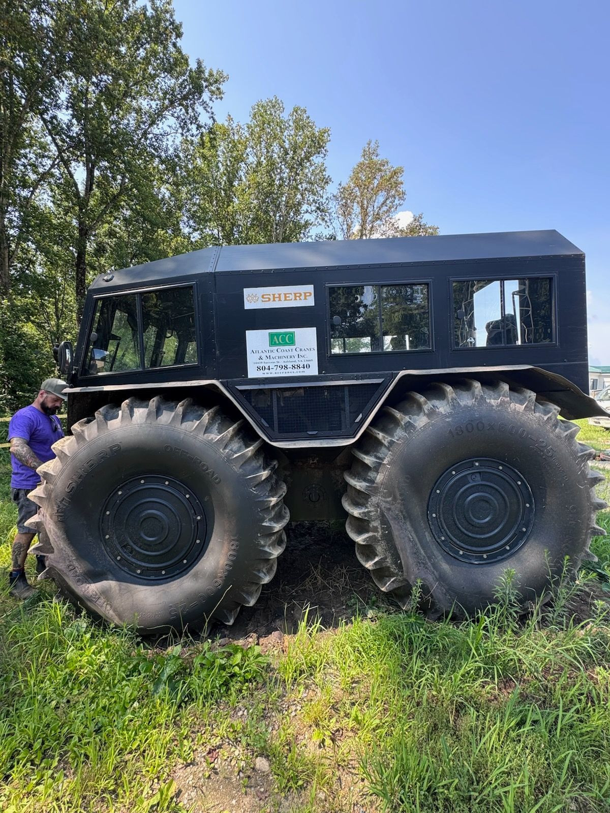 Large black all-terrain vehicle with oversized tires parked on grass, next to a person.
