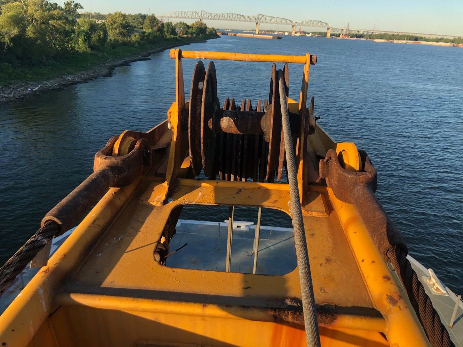 Yellow barge deck with metal rigging, looking out onto a river with a bridge in the distance.