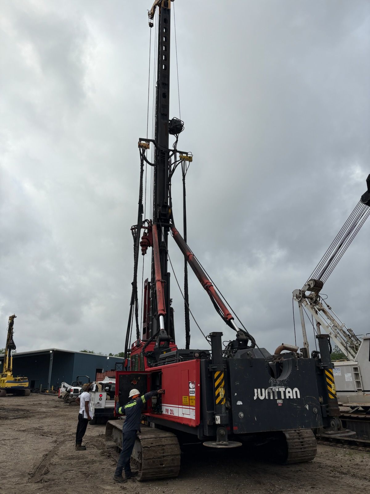 Red and black drilling rig on tracks; workers beside it, cloudy sky.