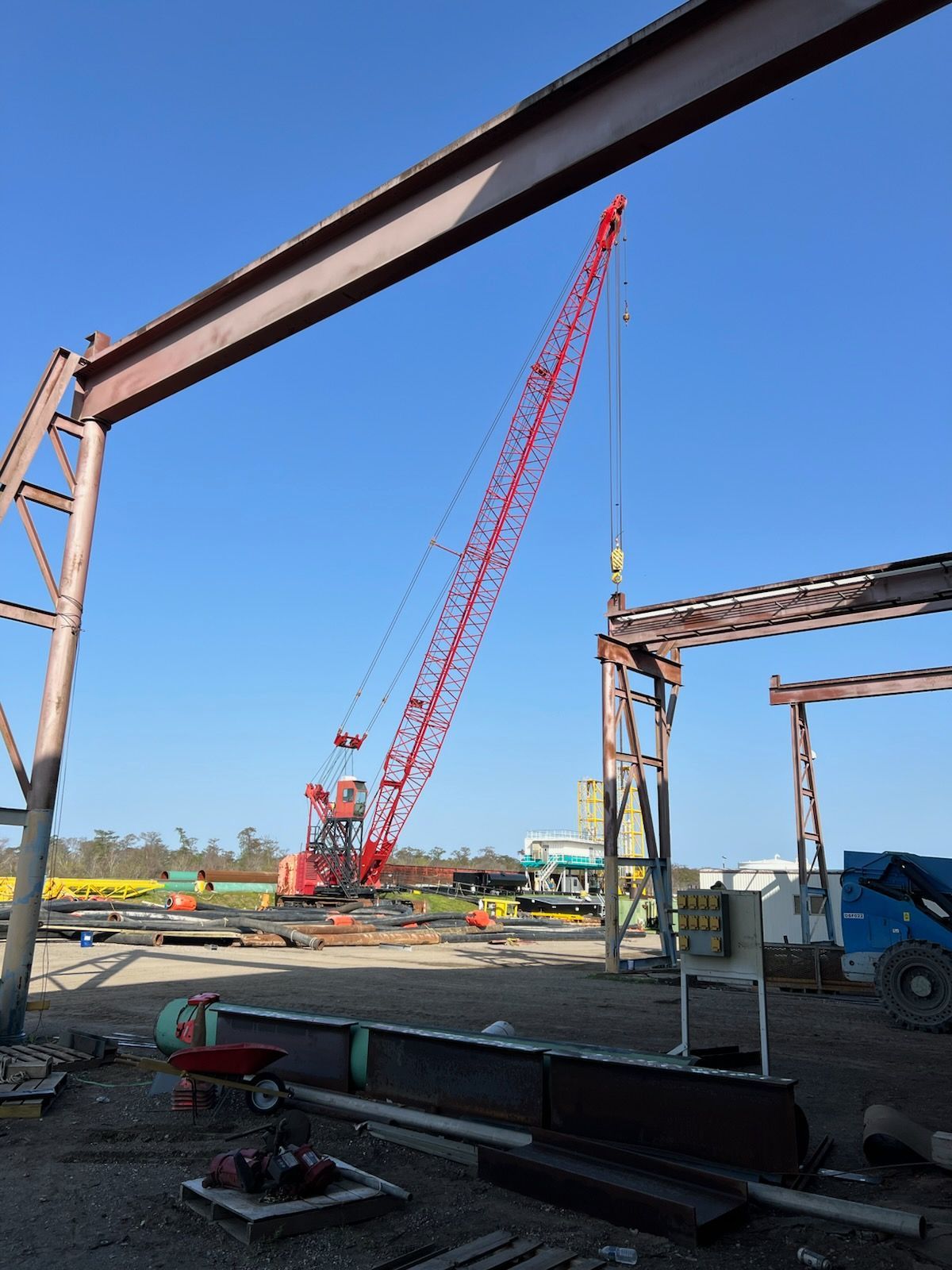A large red crane lifting a steel beam in an outdoor construction site, with a blue sky.