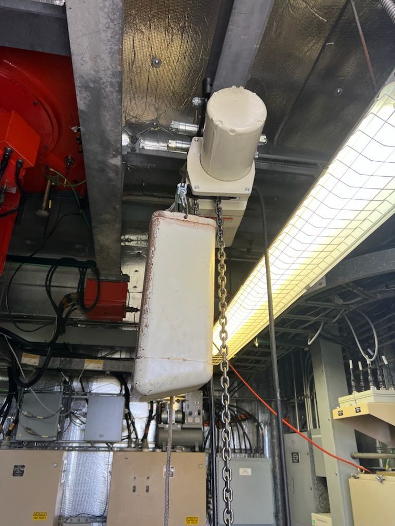 Overhead view of equipment: a white rectangular object suspended by a chain from a motorized hoist in a metallic, industrial setting.