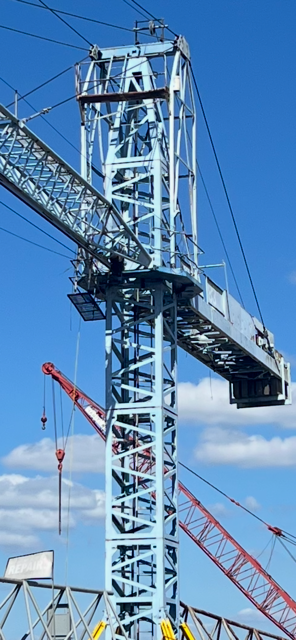 A tall blue construction crane against a blue sky with some clouds. Another red crane is in the background.
