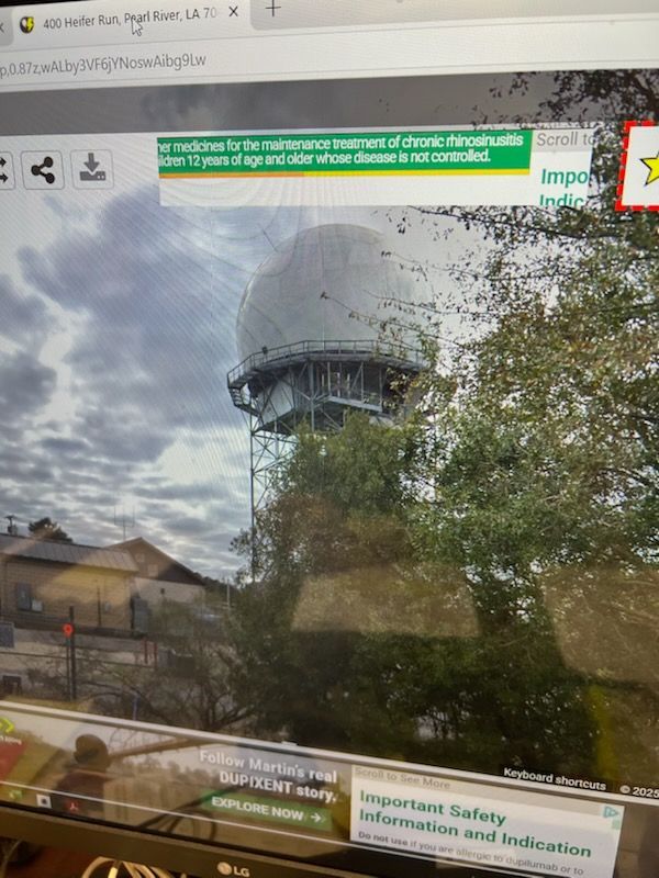 Radar dome on a tower behind trees and a building against a cloudy sky.