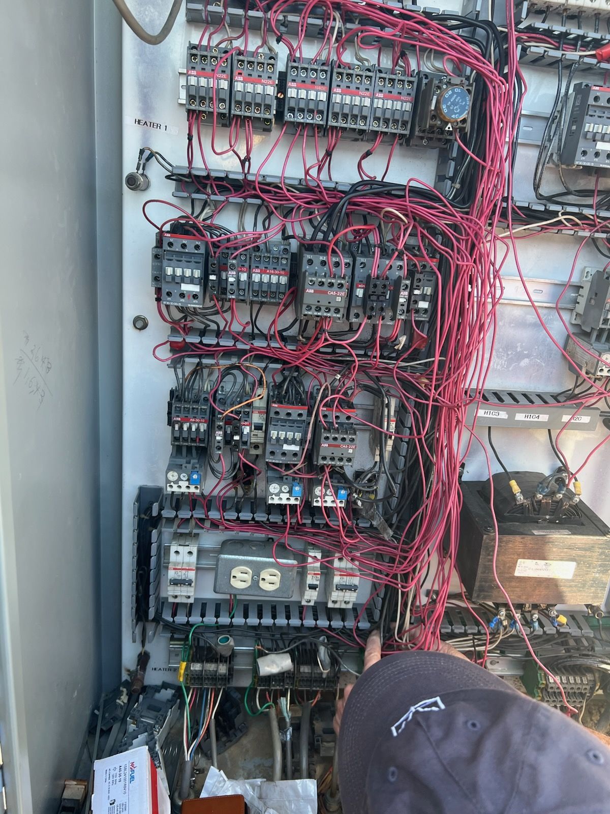 Electrical control panel with complex wiring, red wires dominate. Person points at bottom.