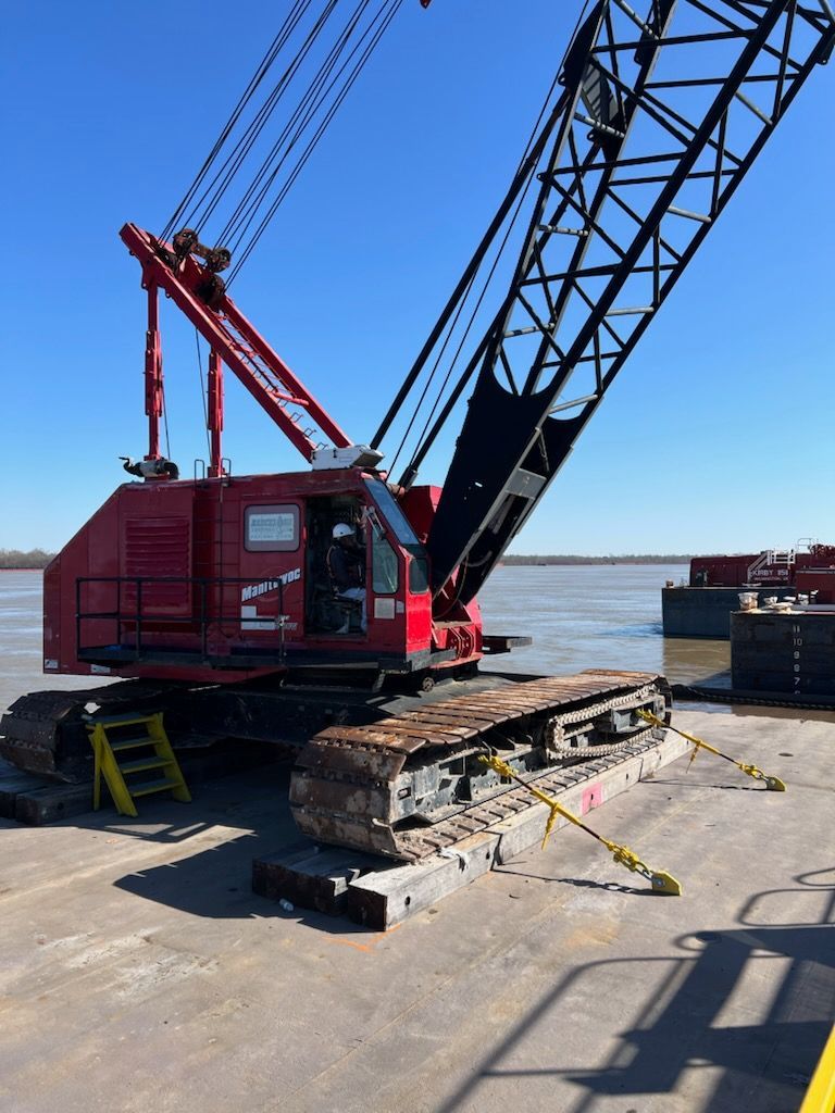 Red crawler crane on a barge, supporting its tracks with yellow straps. Blue sky and water in background.
