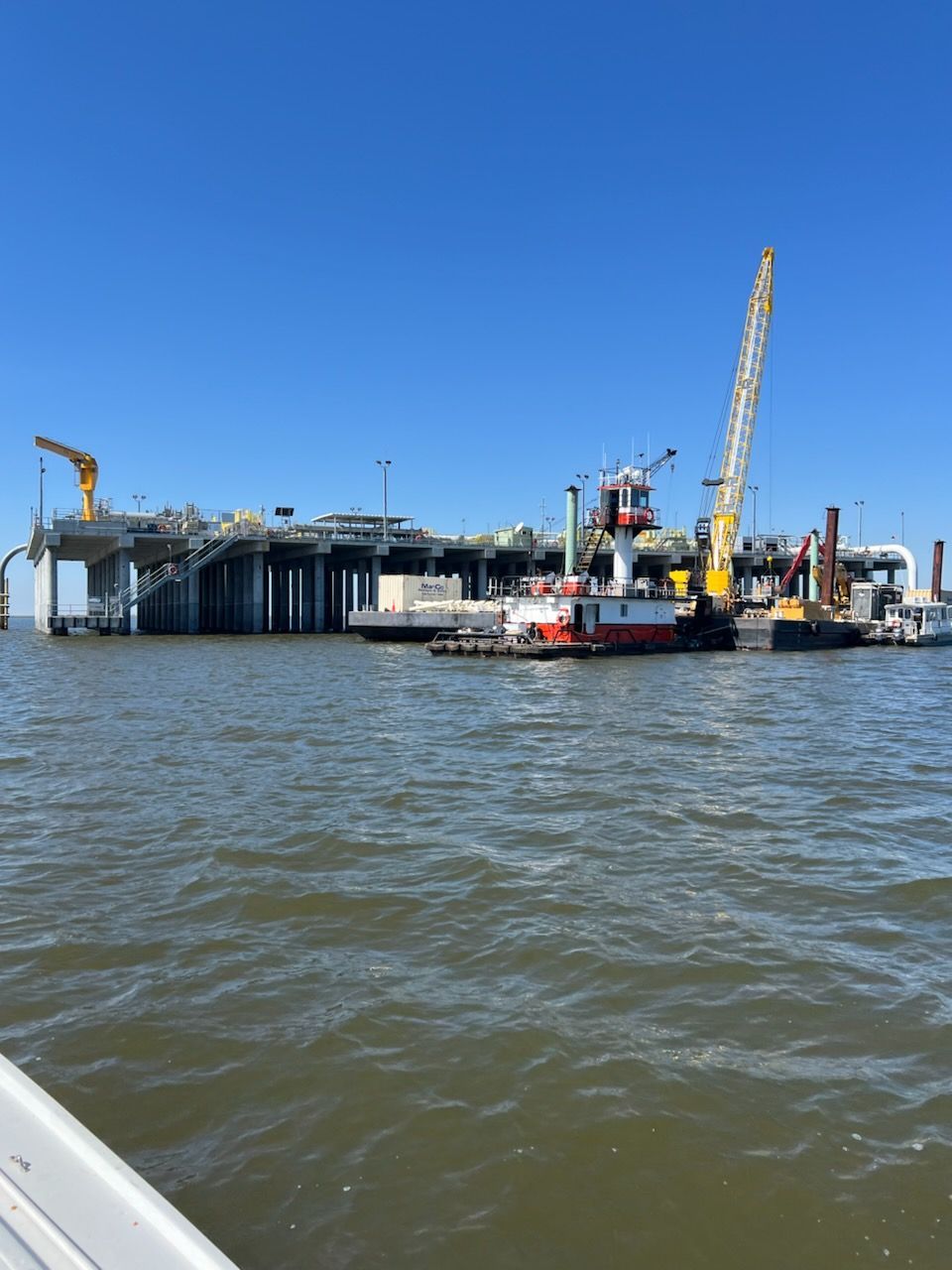 Oil platform with support pilings, cranes, and tugboats in blue water under a clear sky.