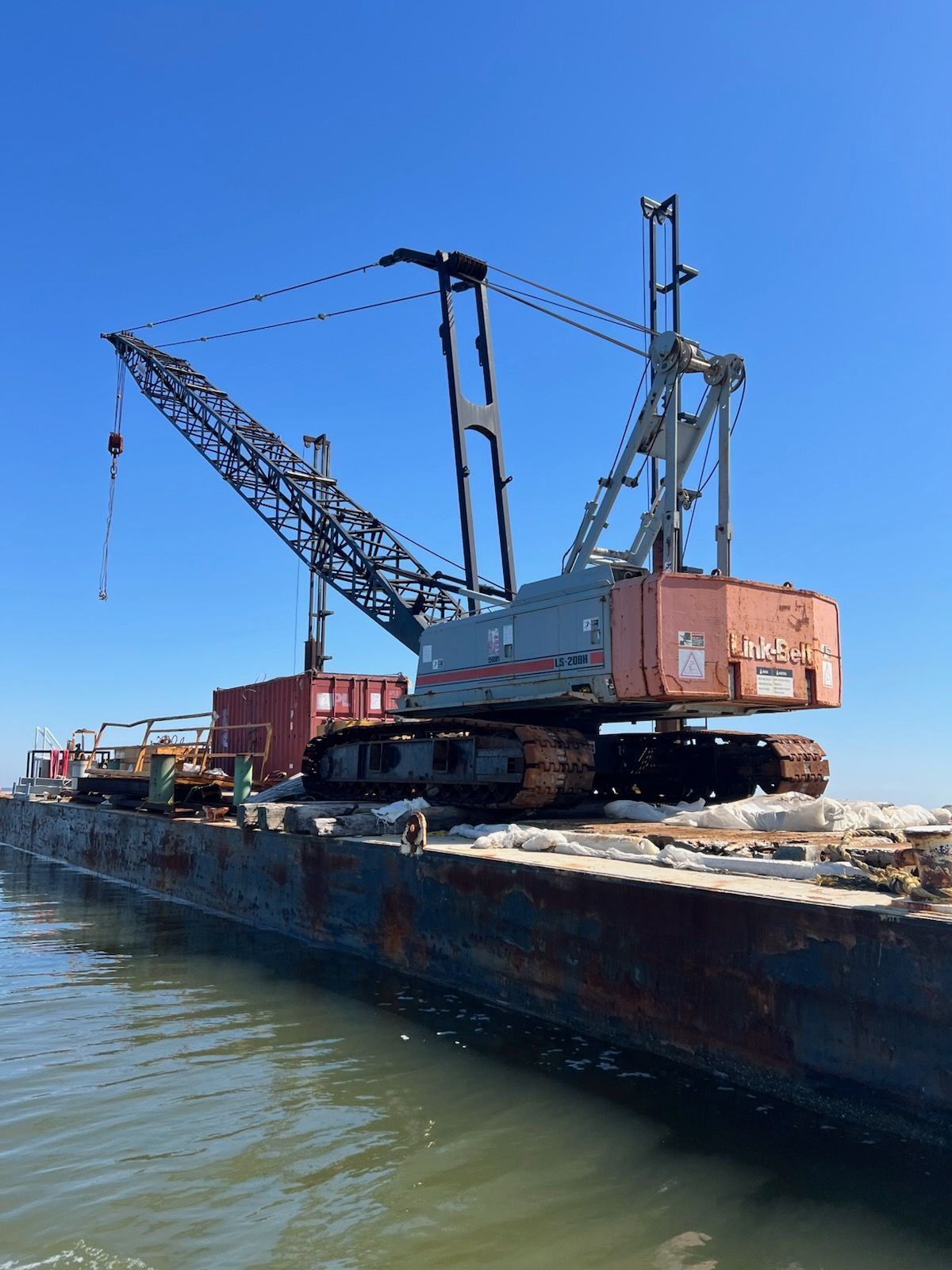 Crane on a barge in water under a blue sky; brown and red construction equipment.
