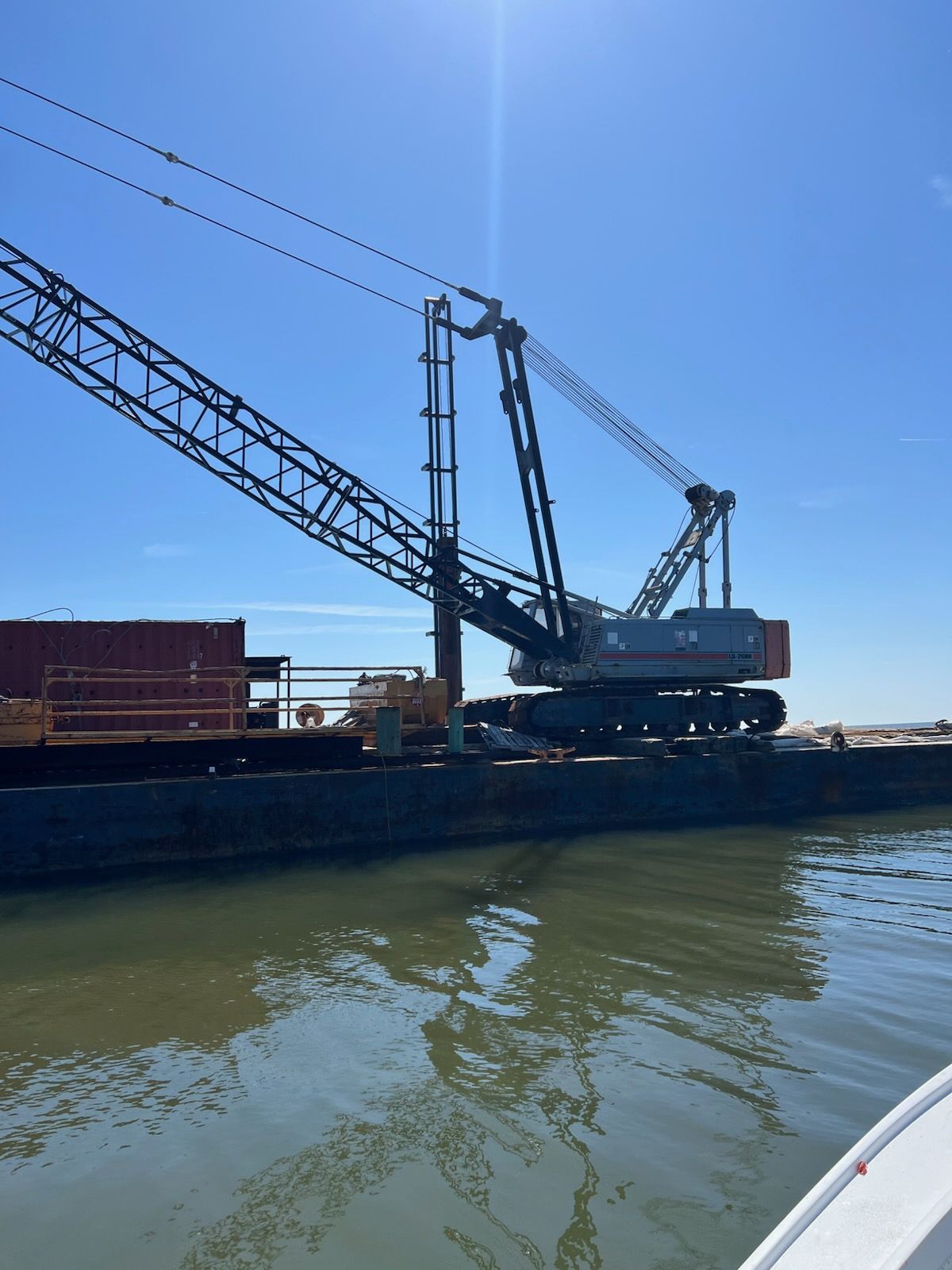 Crane on a barge in murky water under a bright sky.
