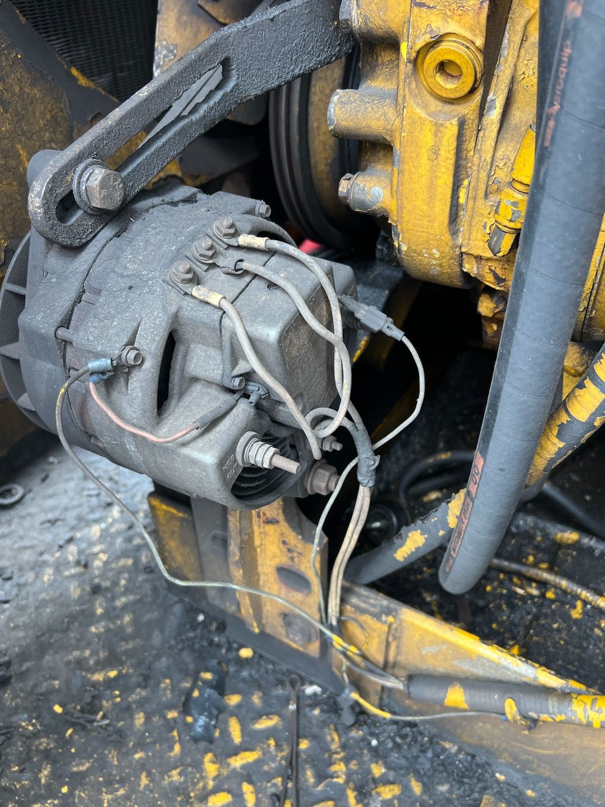 Alternator with connected wires, mounted on a yellow engine, covered in debris.