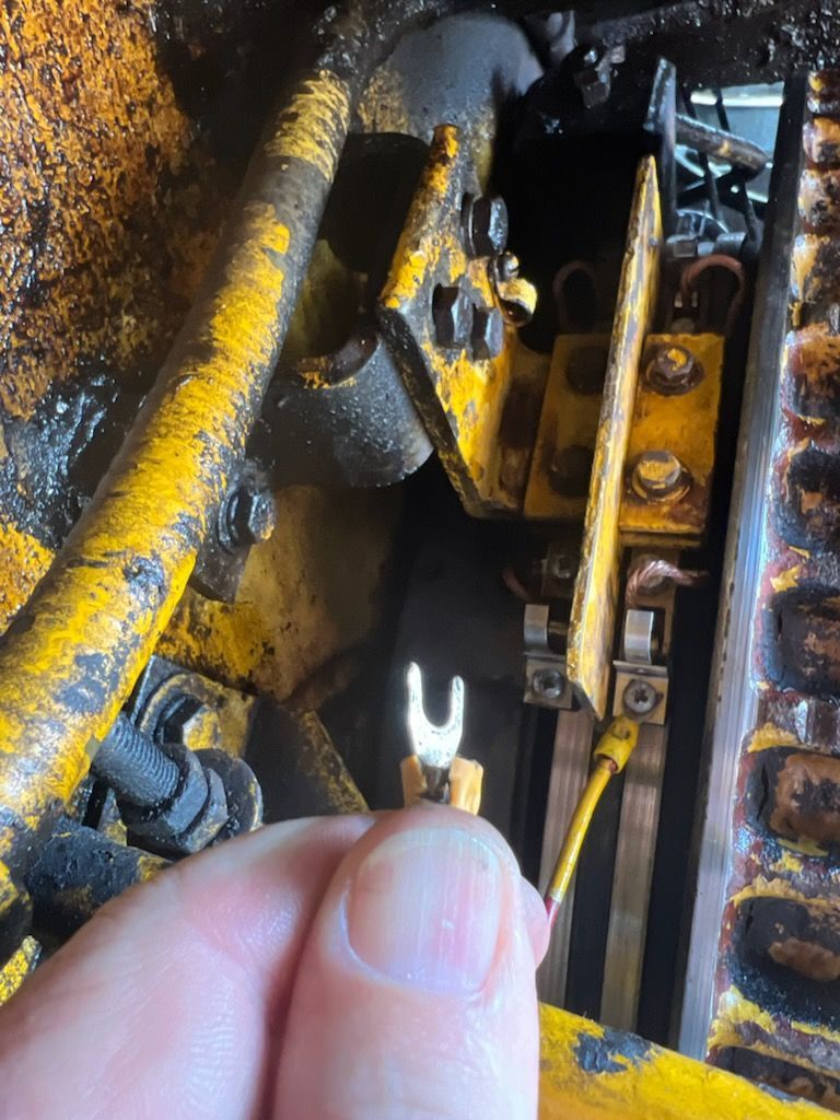Hand holding a U-shaped electrical connector near yellow machinery with bolts and wires.