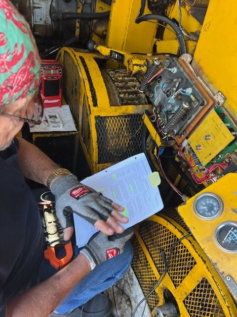 Person examining machinery, reading paperwork, wearing gloves. Yellow equipment, gauges, and electrical components visible.