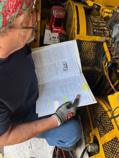 Man with glasses and a headscarf reads a manual while working on a large yellow machine.