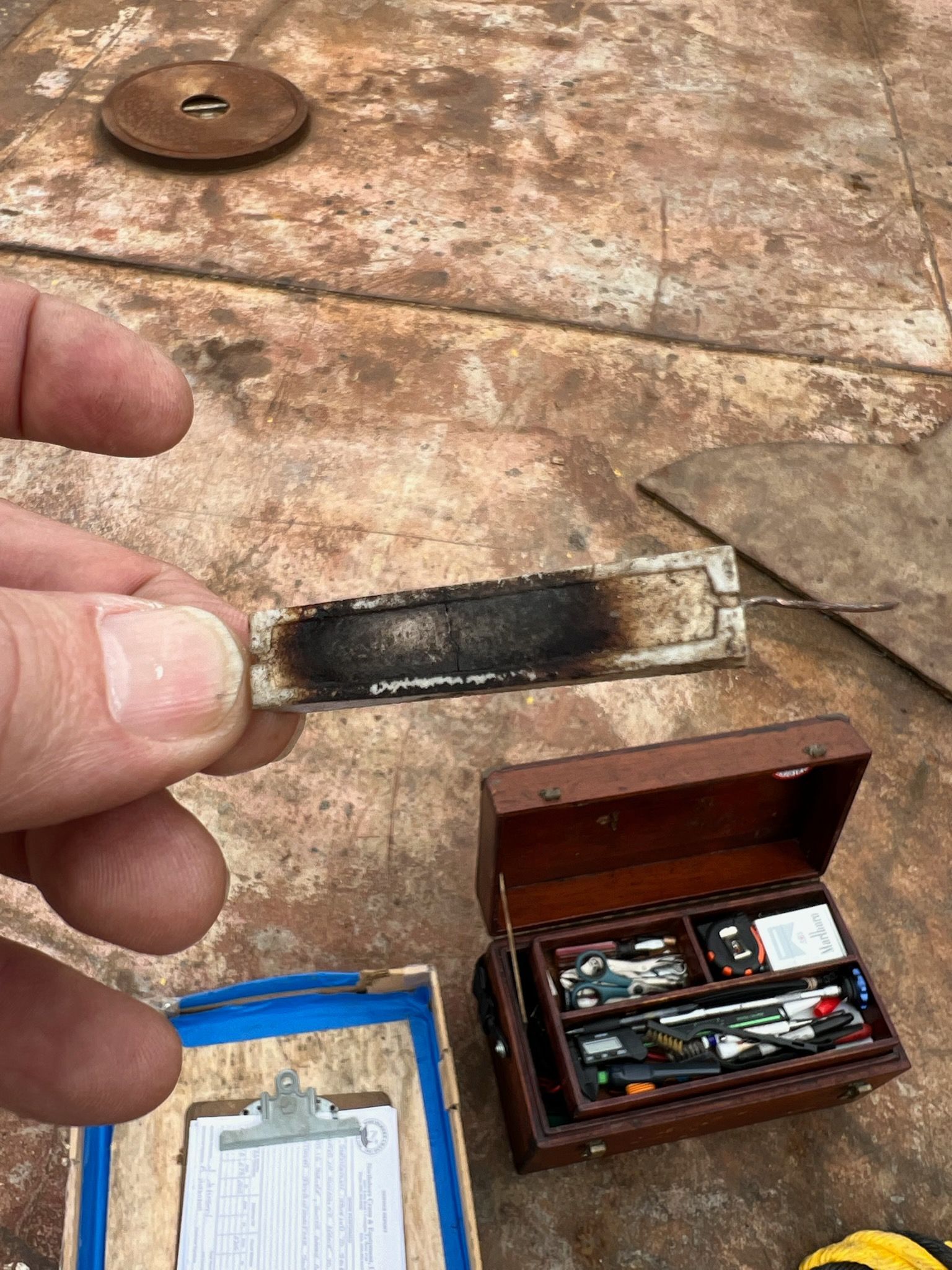 Hand holding a severely burnt electronic component. Small toolbox and clipboard in the background.