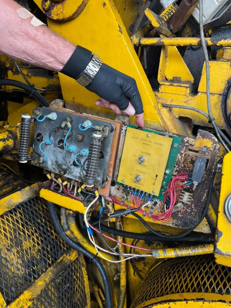 Person's gloved hand points at an exposed circuit board within a yellow machine. Wires and metal components are visible.