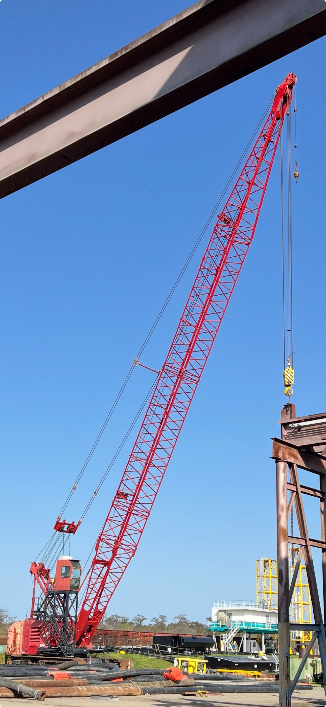 A large red crane lifts a beam at a construction site against a clear blue sky.