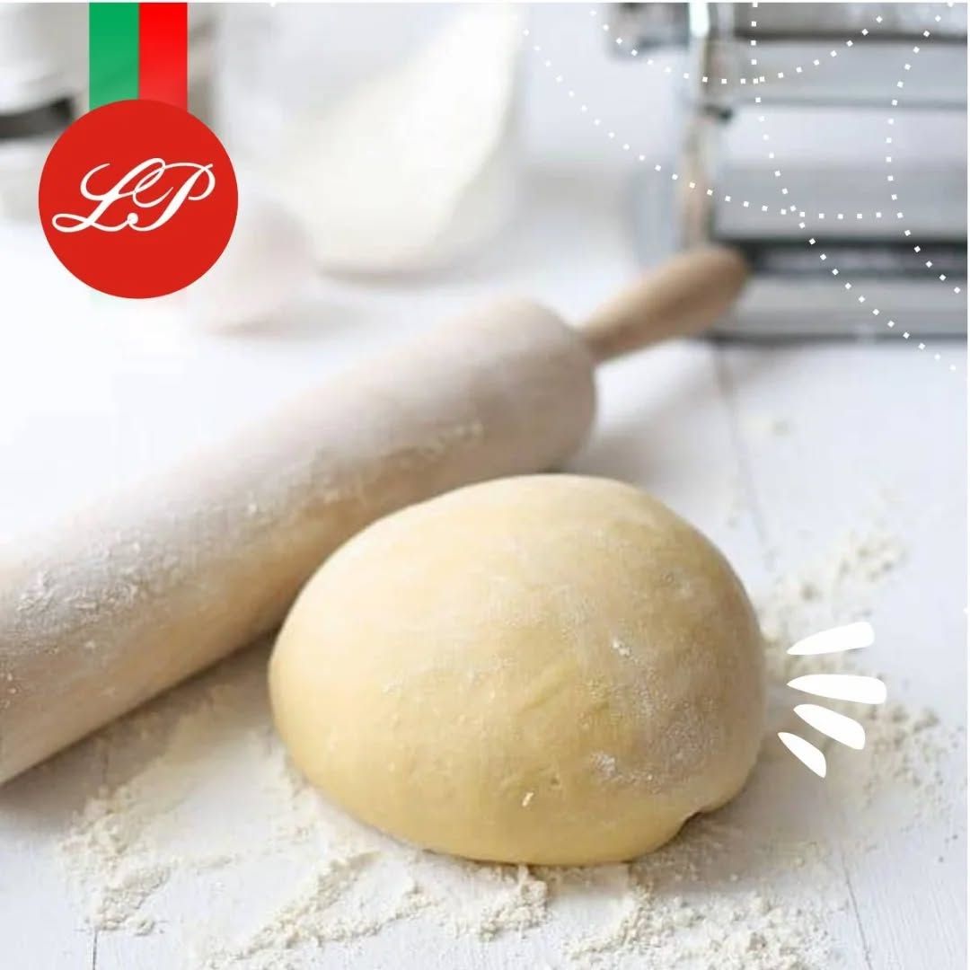 Bola de masa y rodillo sobre una superficie enharinada, con la máquina para pasta y la harina en el fondo.
