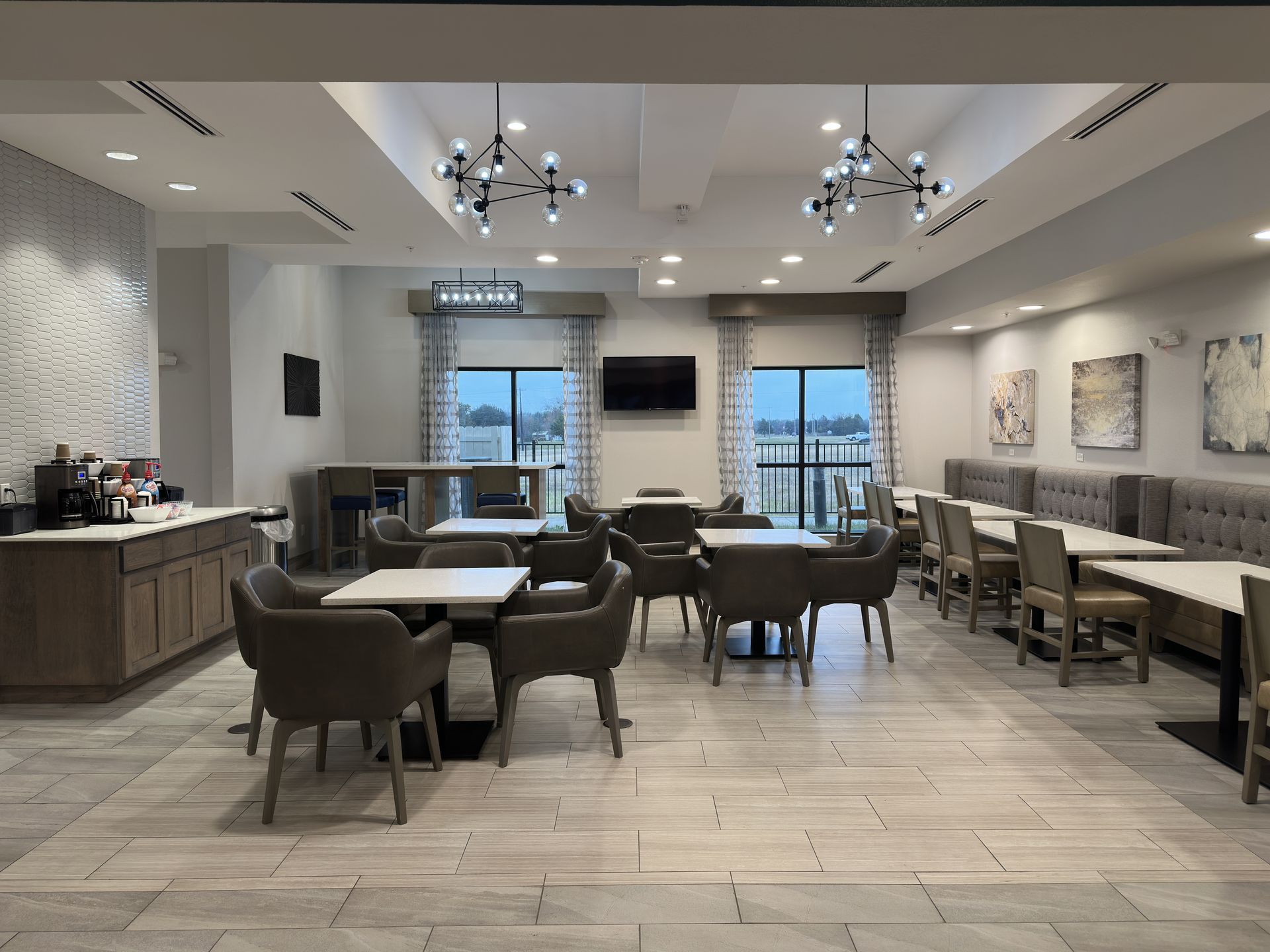 Hotel breakfast area with tables and chairs, coffee station, windows, and decorative lighting.