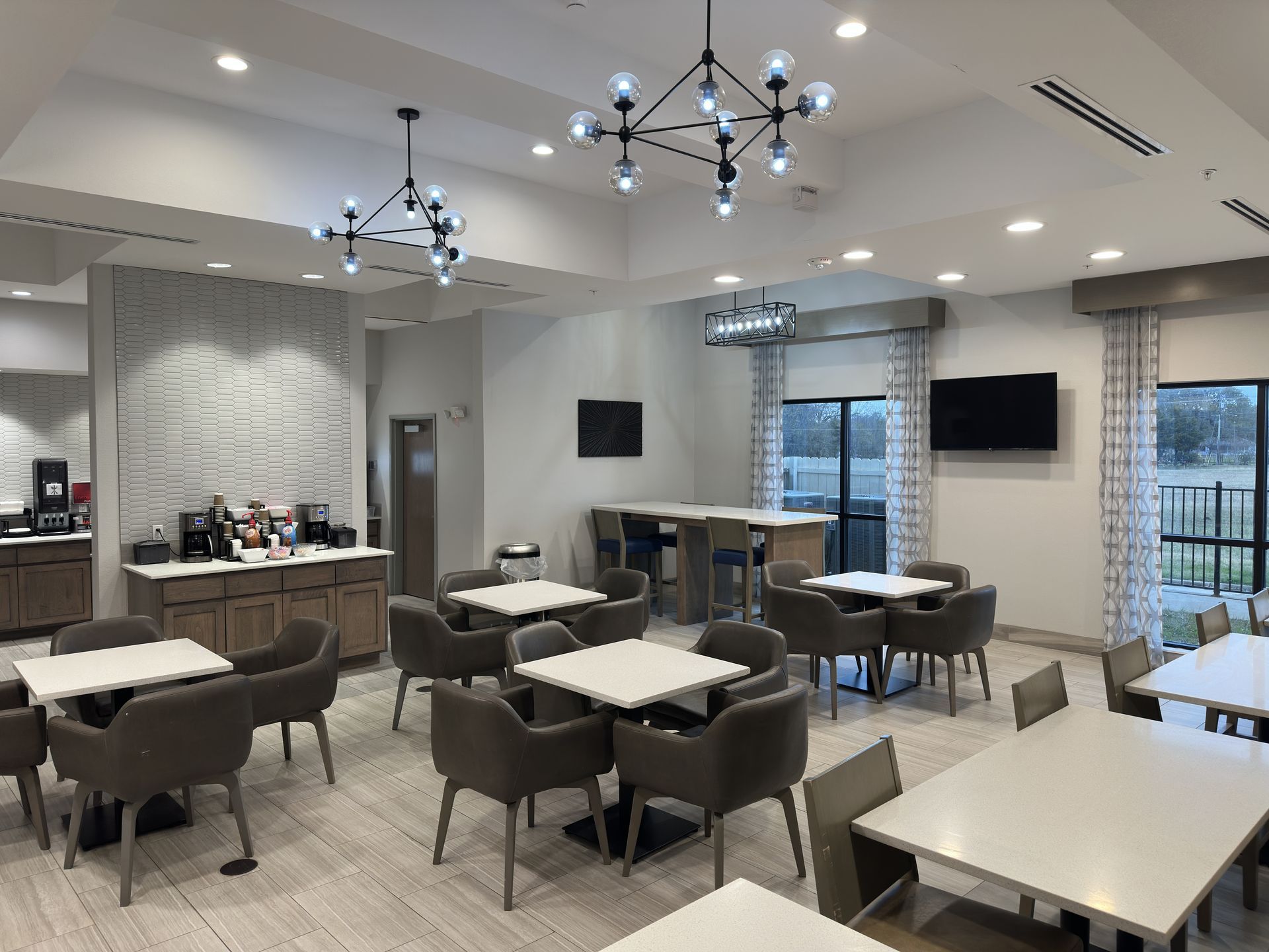 Hotel breakfast area with tables, chairs, coffee station, decorative lighting, and two TVs.