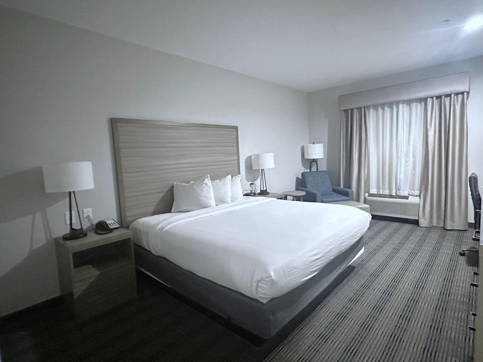 Hotel room with a large bed, two lamps, and a chair near a window with curtains.