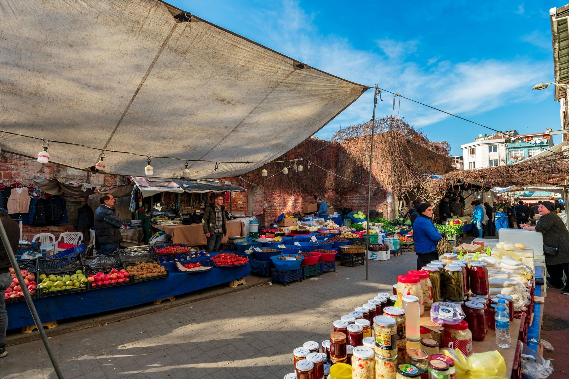 Outdoor market with stalls of produce under a shaded awning. People browse and shop.