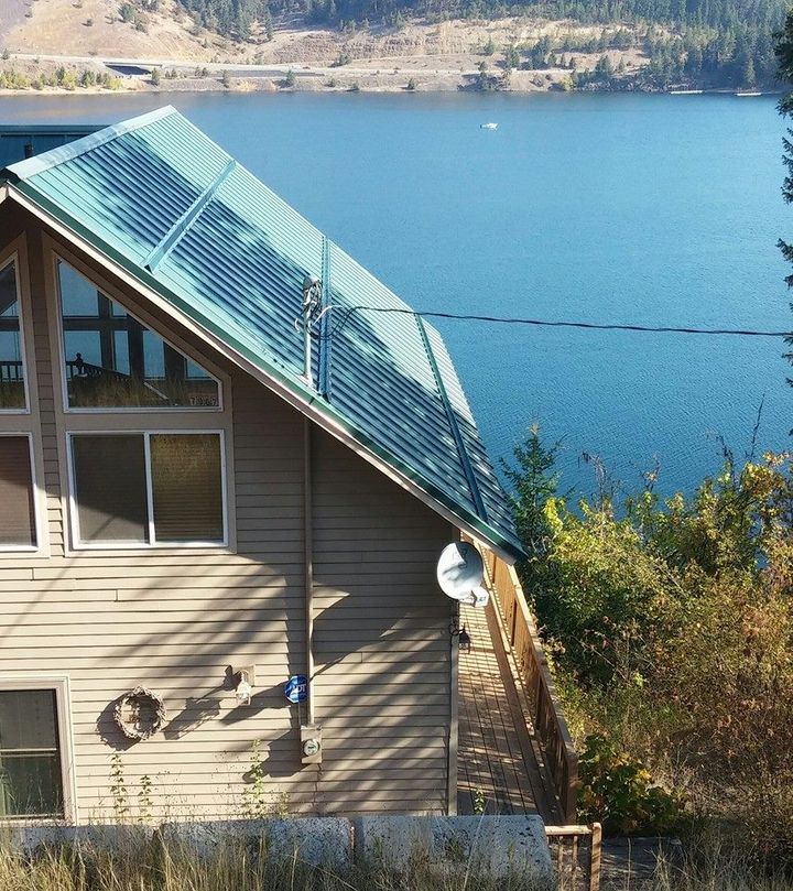 House with green metal roof overlooking a blue lake, with trees and hills in the background