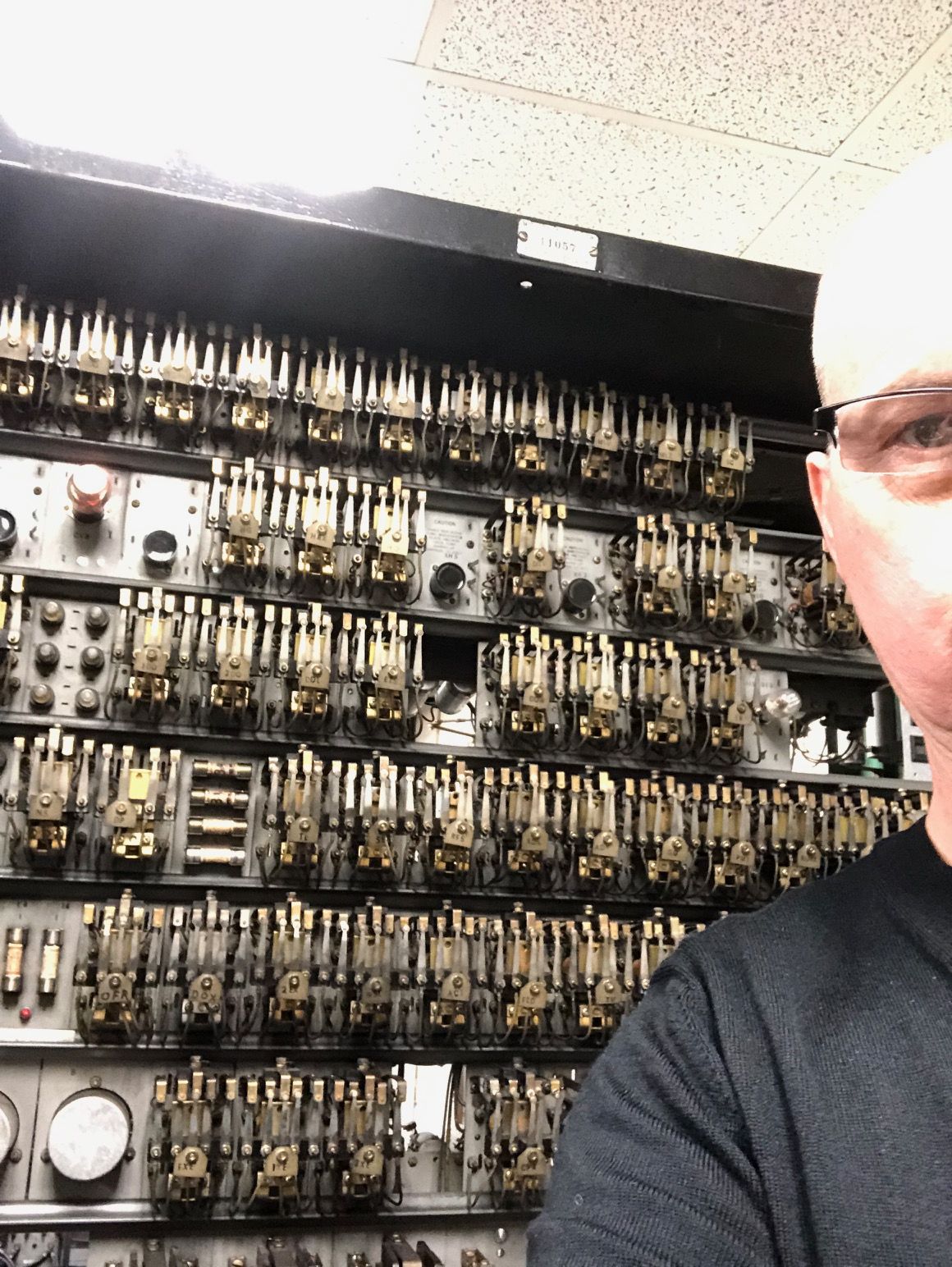 Man with glasses next to a large electrical panel with many relays and gauges.