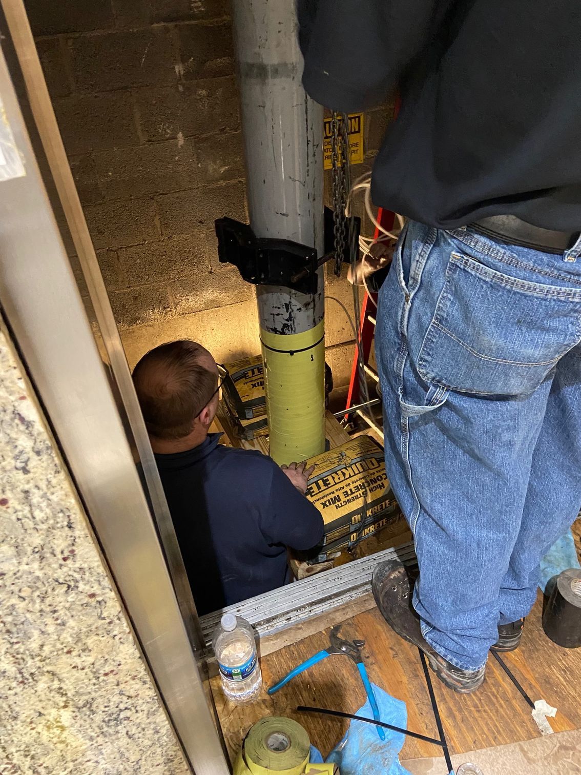 Two workers repair a pipe in a narrow opening; one bends down, the other stands above.