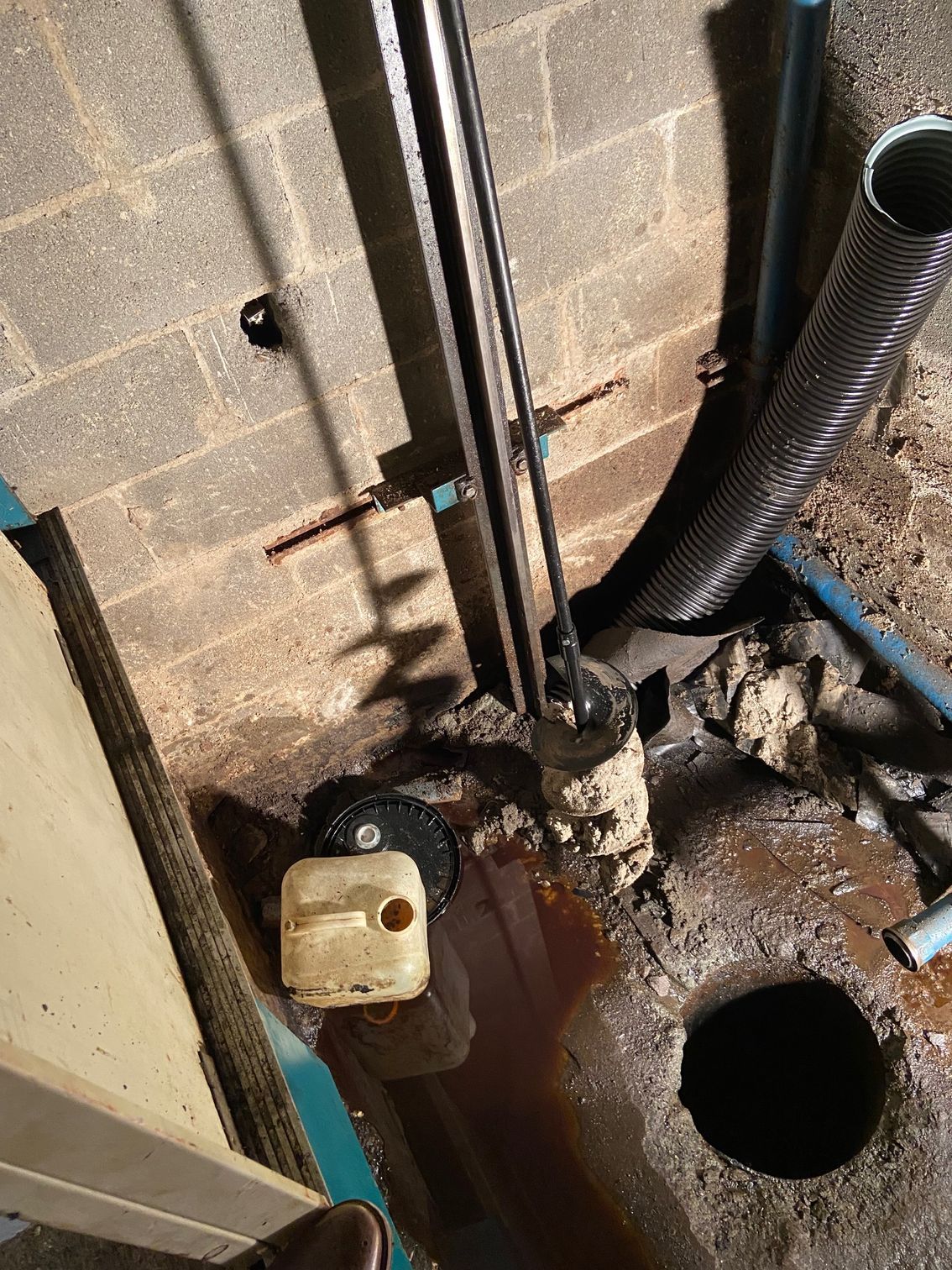 Basement sump pump system, dark brown water, concrete floor, black drain hole, white and black pipes.