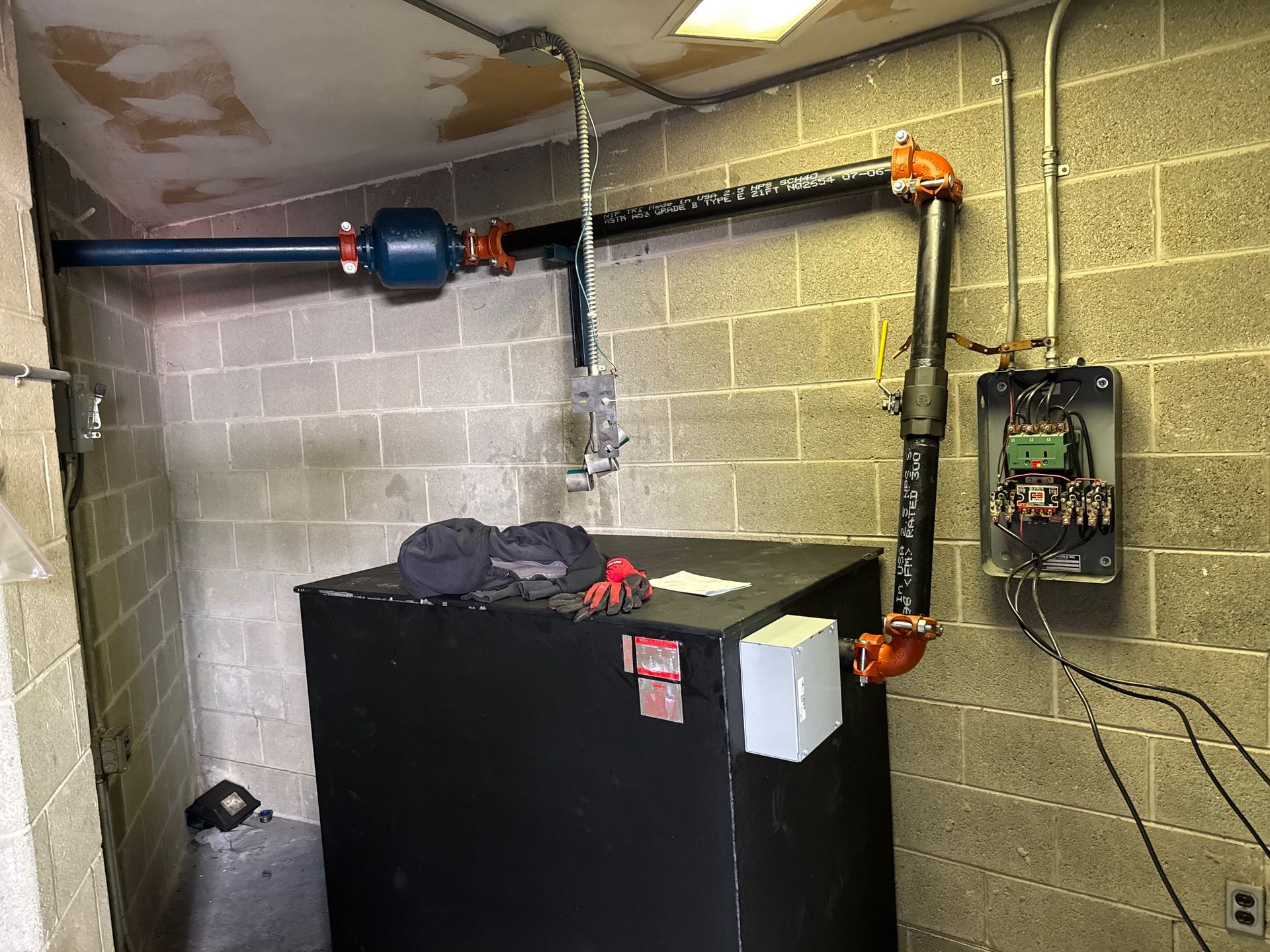 A water tank with piping and electrical box against a cinder block wall in a utility room.