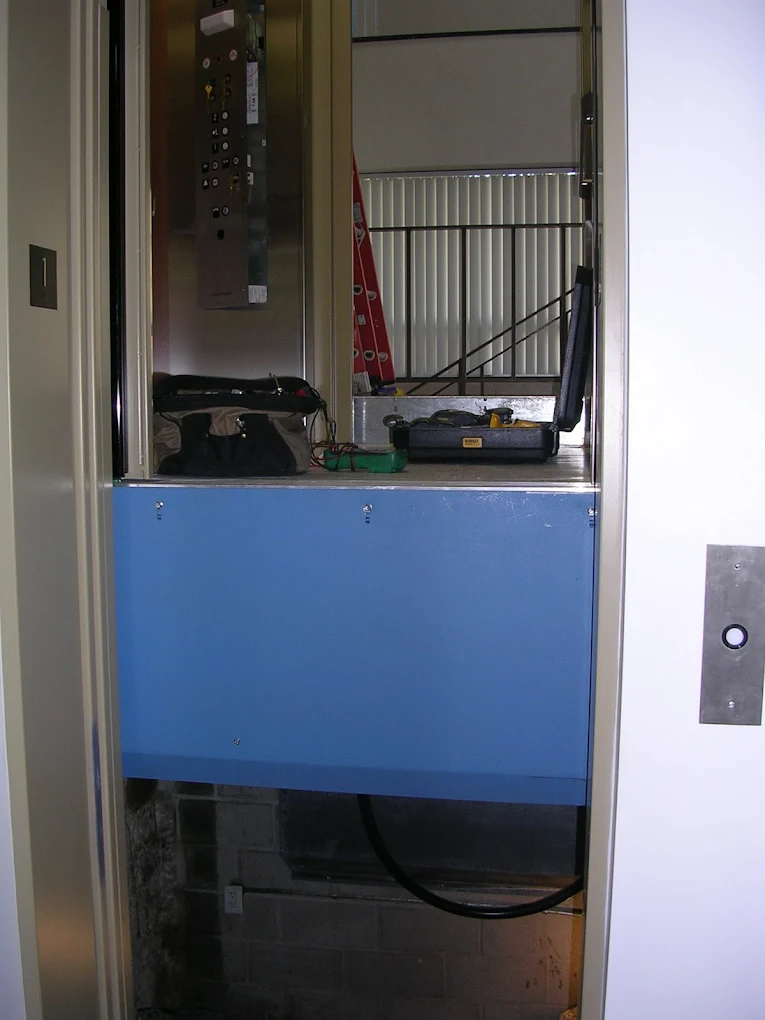 Blue elevator platform inside a shaft. Tools and control panel visible. Doorway on the right.