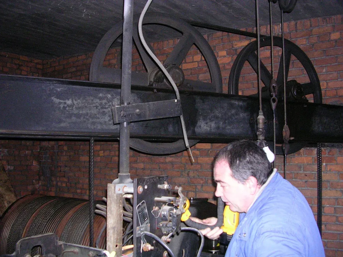 Man in blue examines machinery with large black wheels in brick-lined room.