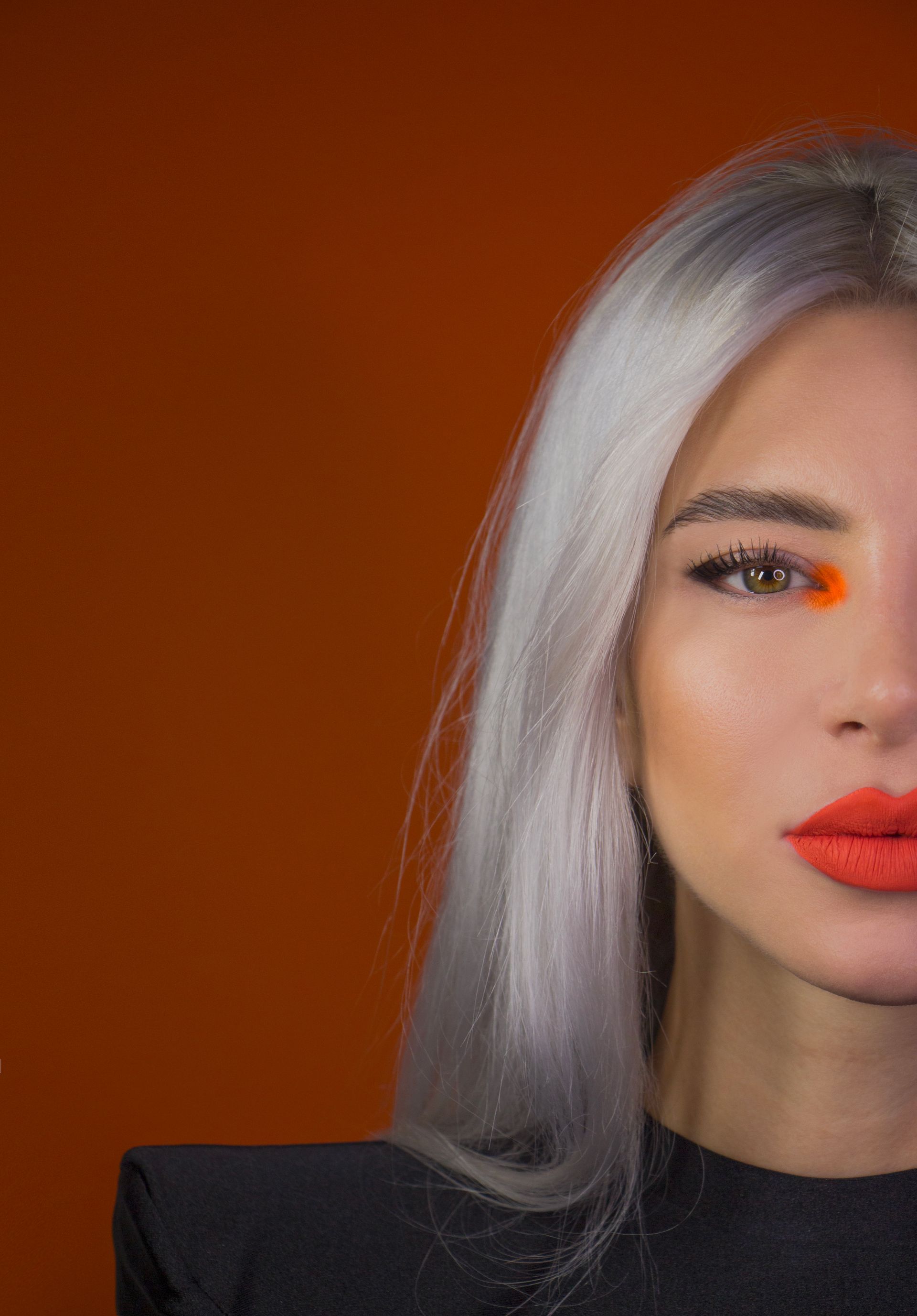 Primo piano di una donna con capelli argentati e rossetto arancione su uno sfondo arancione.