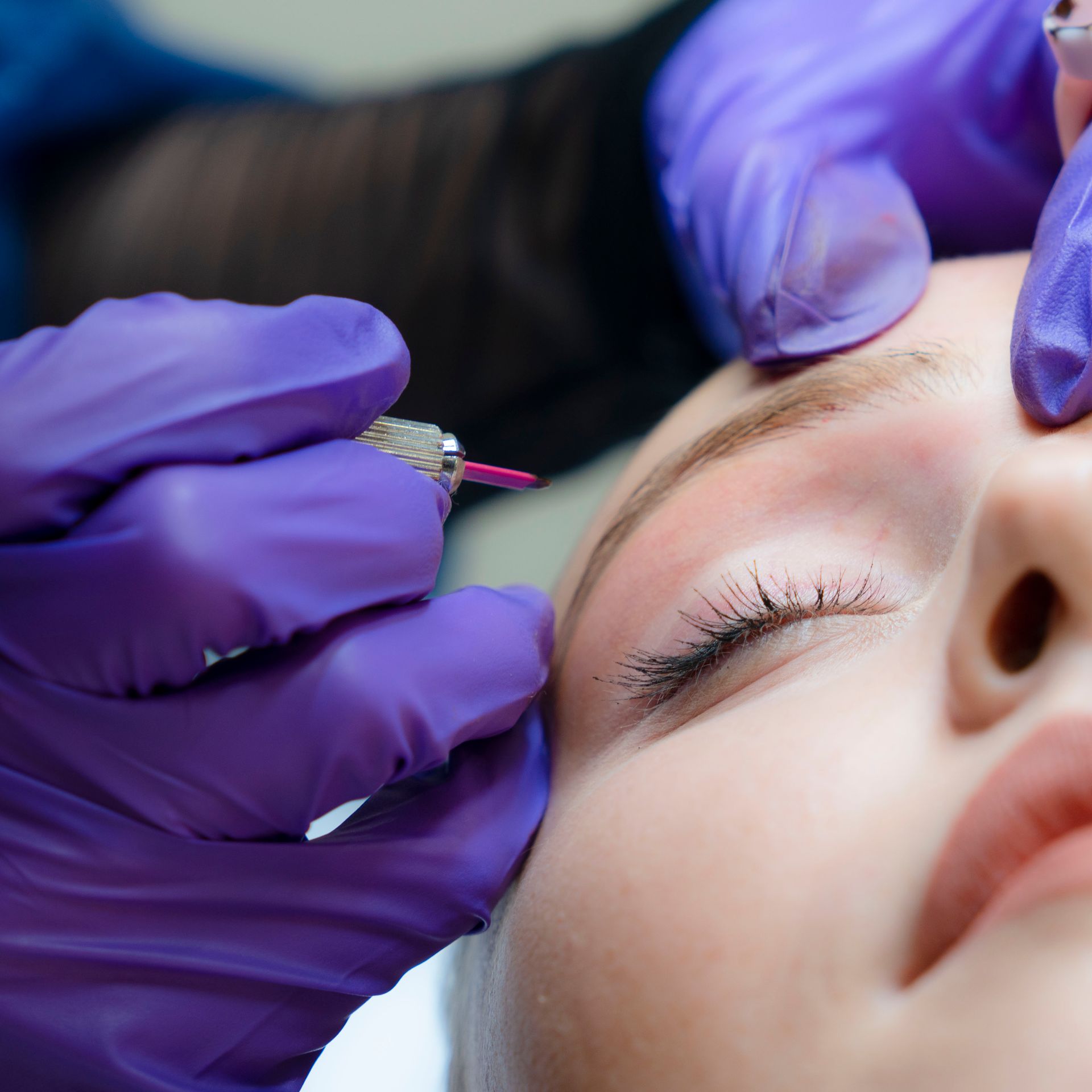 Mani guantate applicano un trattamento cosmetico al sopracciglio di una donna con uno strumento sottile (primo piano).