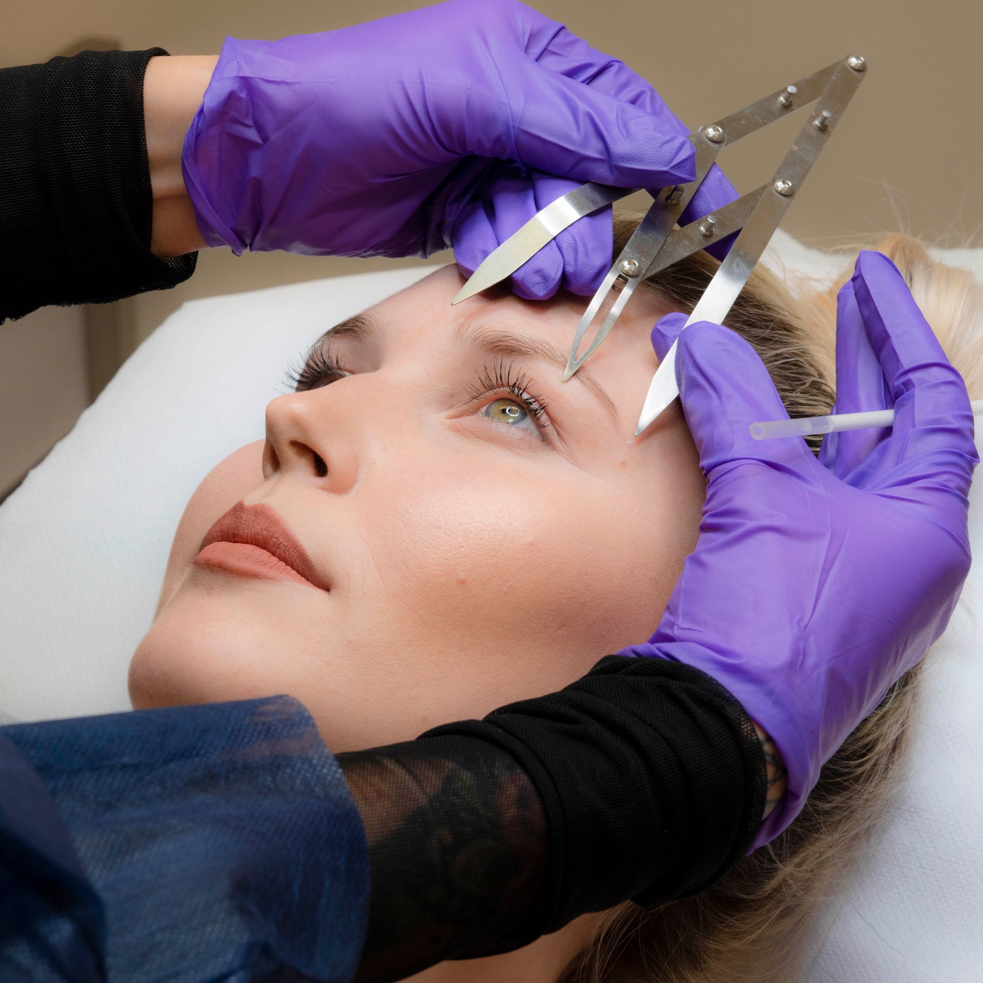 Procedura estetica eseguita con mani guantate e utilizzando un calibro metallico sulla zona delle sopracciglia di una donna.