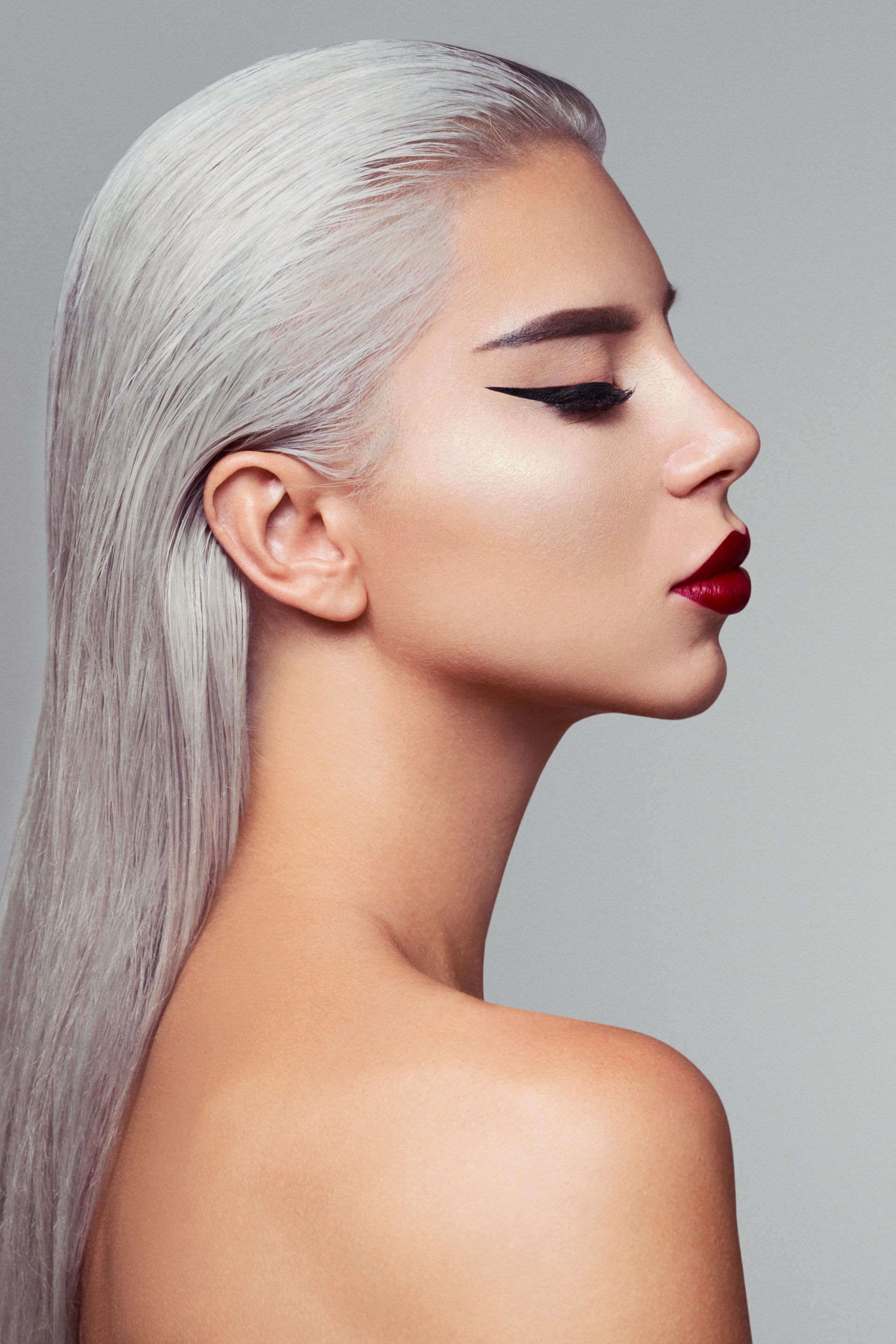 Ritratto di profilo di una persona con capelli biondo platino, eyeliner marcato e rossetto rosso su sfondo grigio.