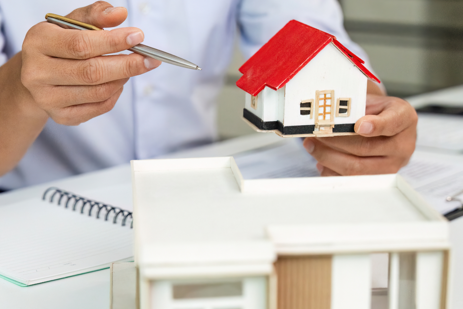 Person holding a model house with red roof, pointing with pen. A second, larger model house is on a table.