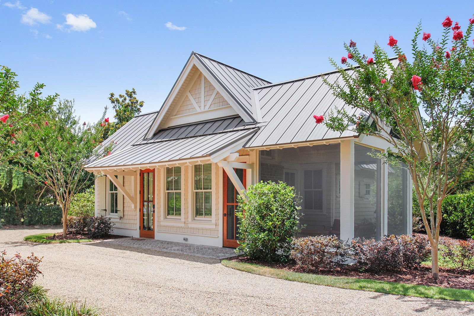 Small white cottage with metal roof, screened porch, and gravel driveway. Lush greenery surrounds the building.