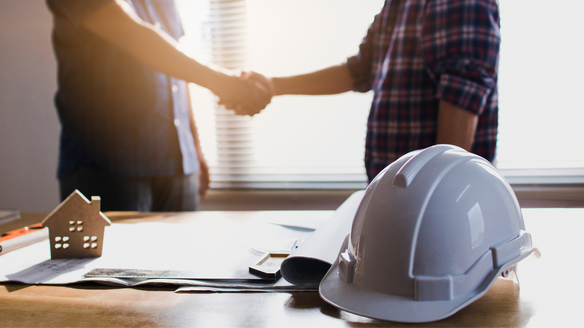 Two people shaking hands over a table with a model house, blueprints, and a hard hat.