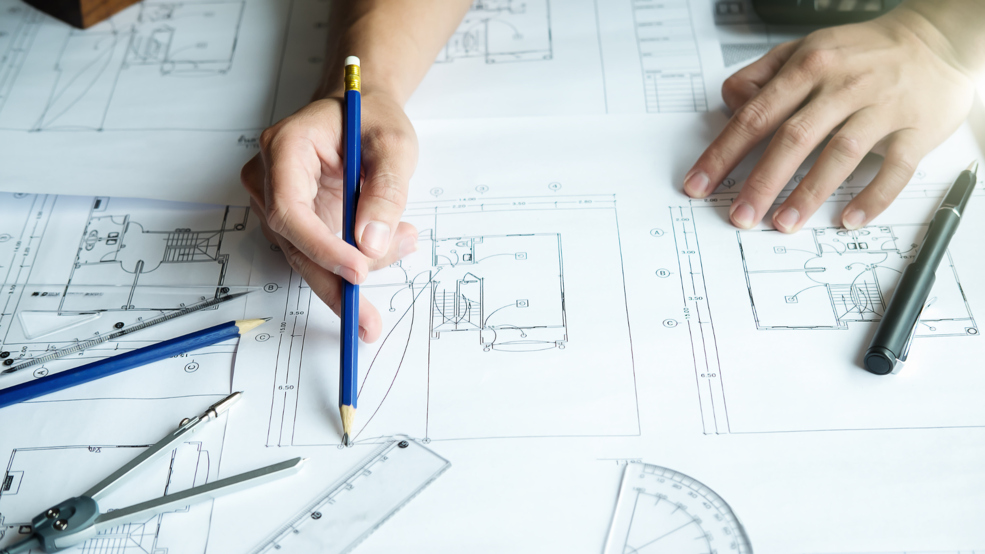 Person's hands drawing on architectural plans with a pencil, desk with drawing tools.
