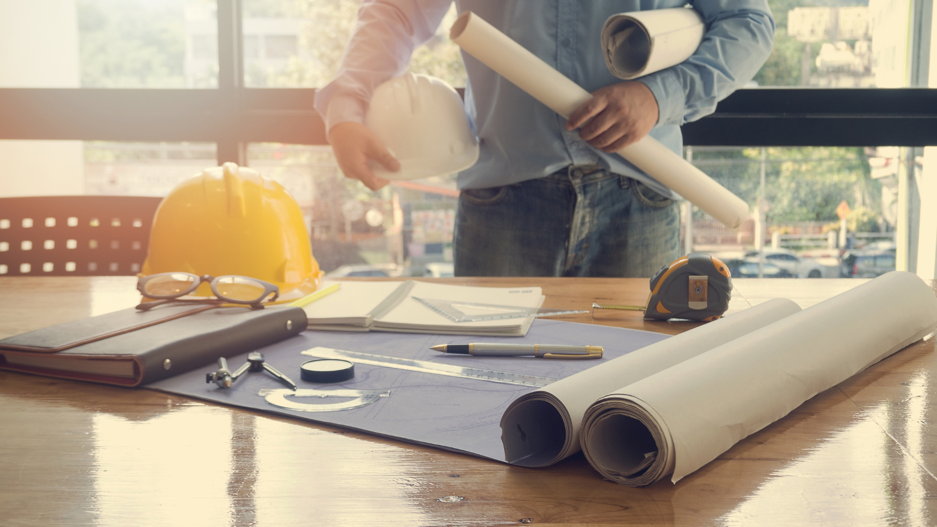 Engineer at a desk with blueprints, holding a hard hat and rolled plans, in a sunny office.