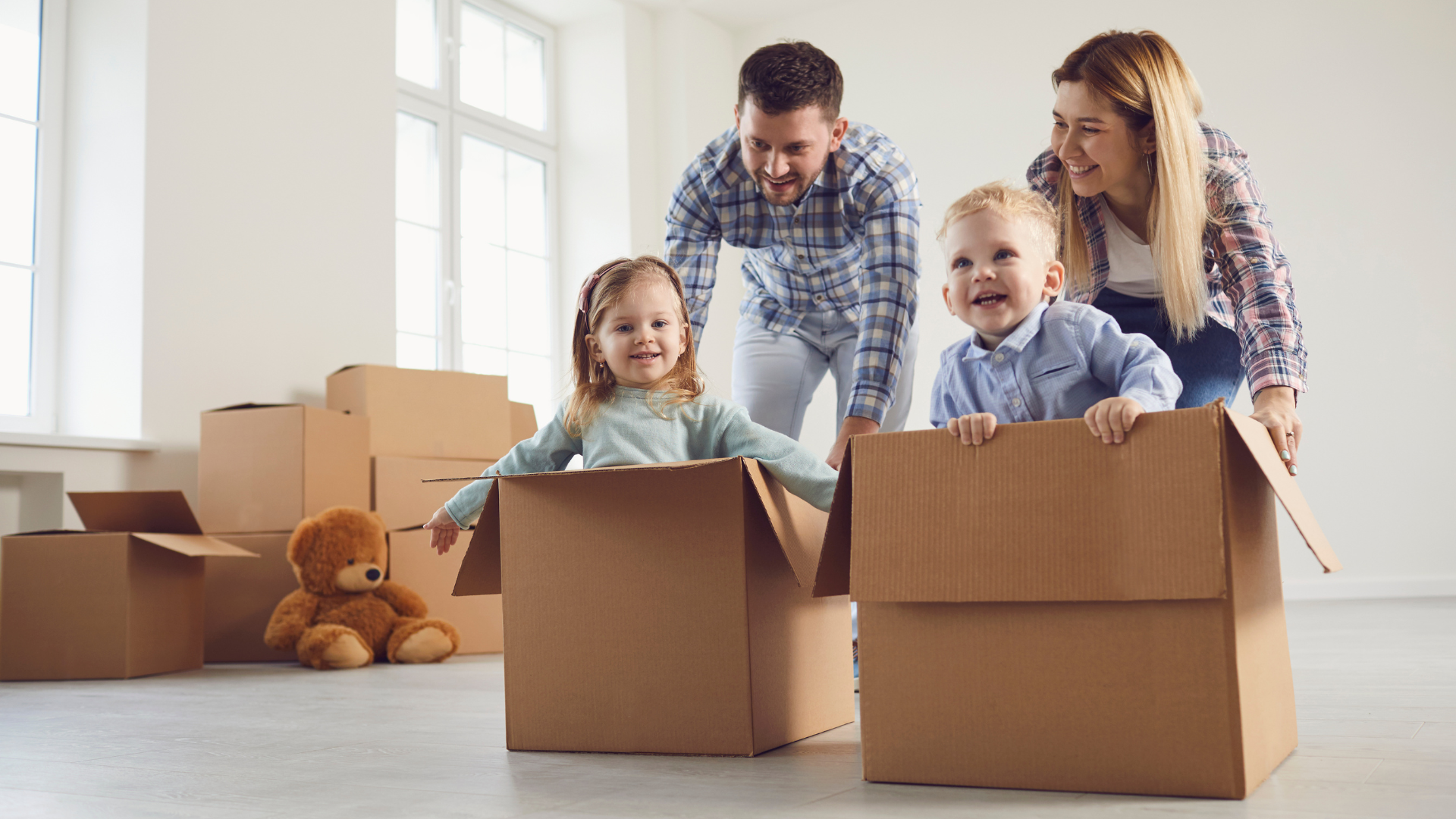 Family in new home, children in boxes, parents smiling. Boxes and teddy bear on the floor.