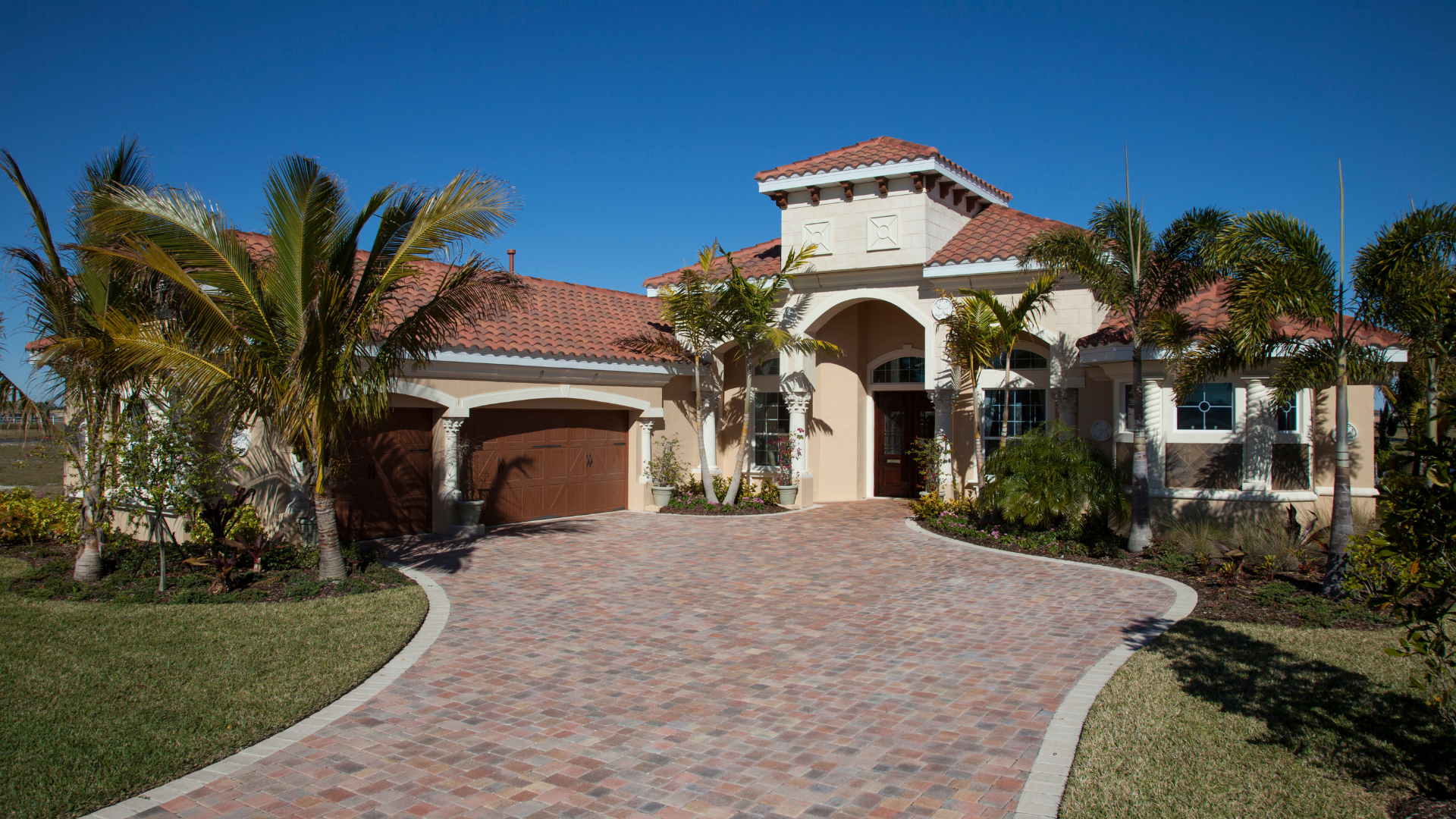 Mediterranean style home with brick driveway, palm trees, and blue sky.