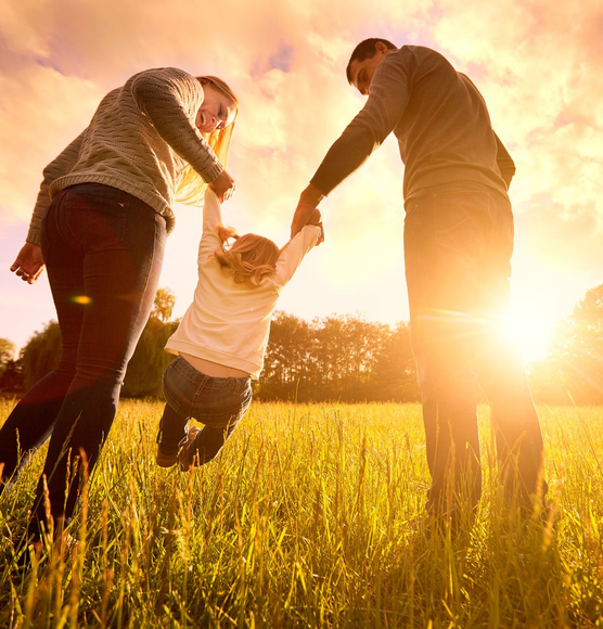 Parents swinging child in a field, silhouetted against the setting sun.