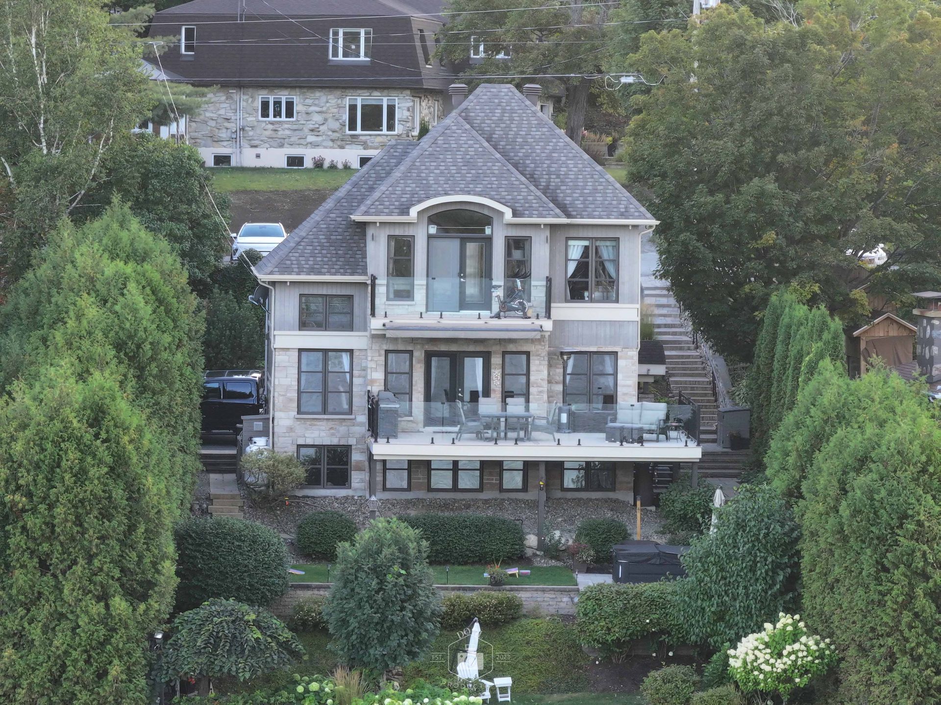 Maison de trois étages avec un toit gris, une façade en pierre et des balcons, entourée d'arbres et donnant sur l'eau.